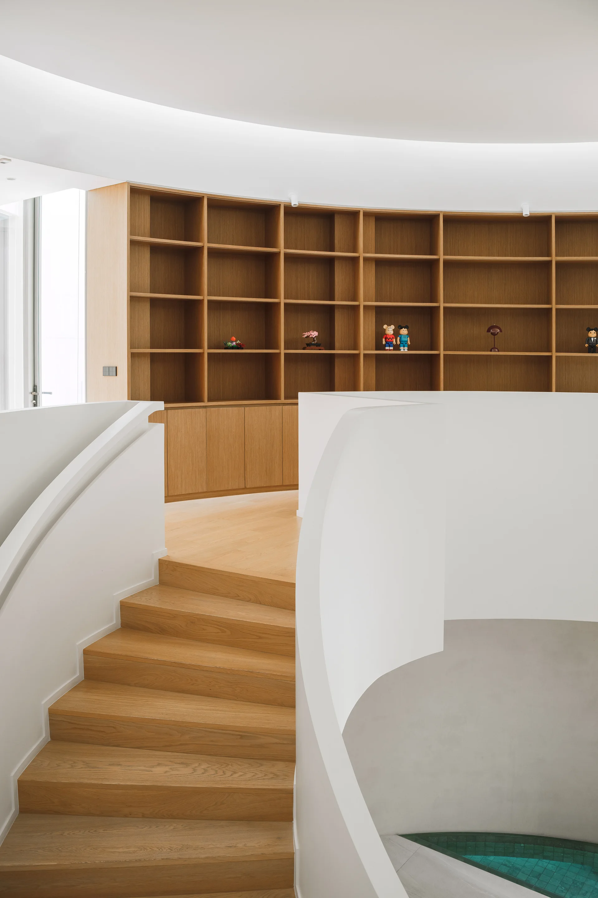 A curved white staircase with light wood steps leads to a landing featuring a large built-in wooden bookcase under soft cove lighting.