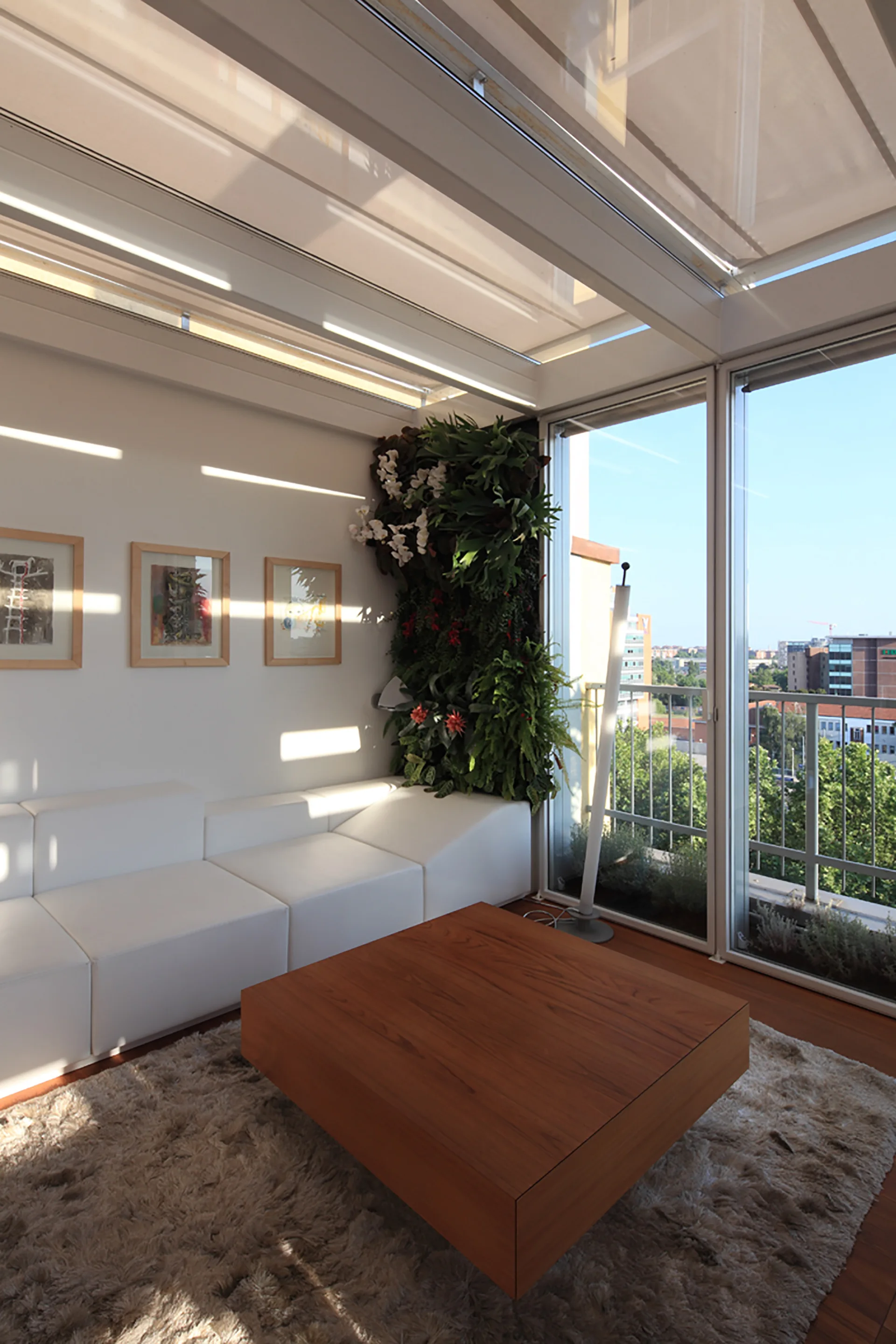 A bright sunroom features a low white modular sofa, a square wood coffee table on a shag rug, and a lush vertical garden next to large windows.