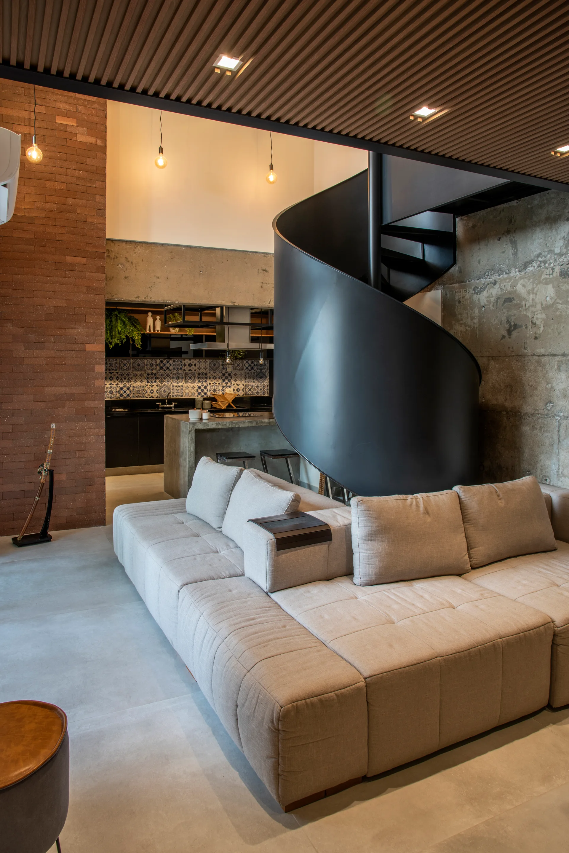 A modern living room featuring a modular beige sofa, a black metal spiral staircase, and an industrial background of concrete and brick walls.