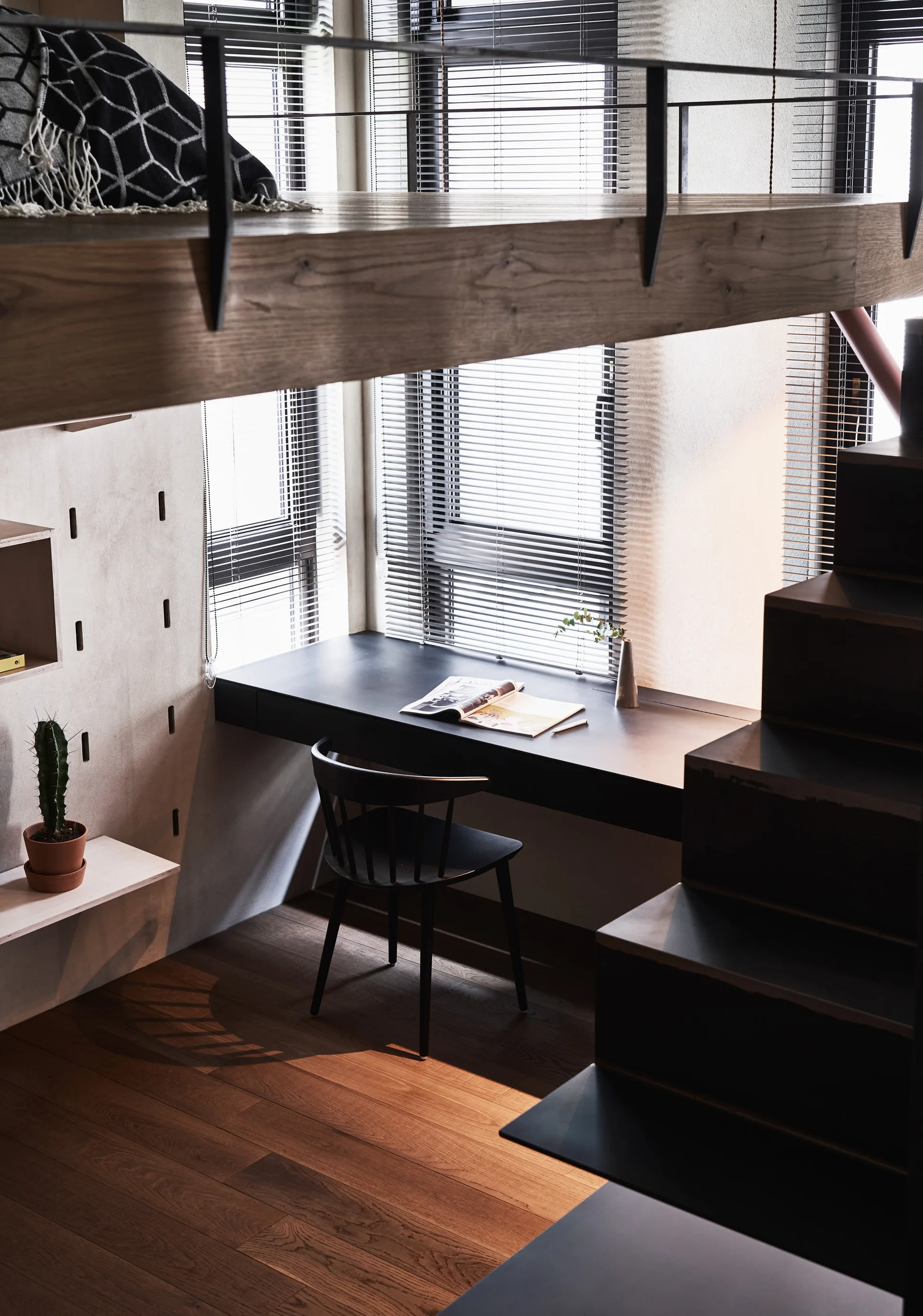 A minimalist home office loft featuring a dark built-in desk beneath tall windows with blinds and a black metal staircase.
