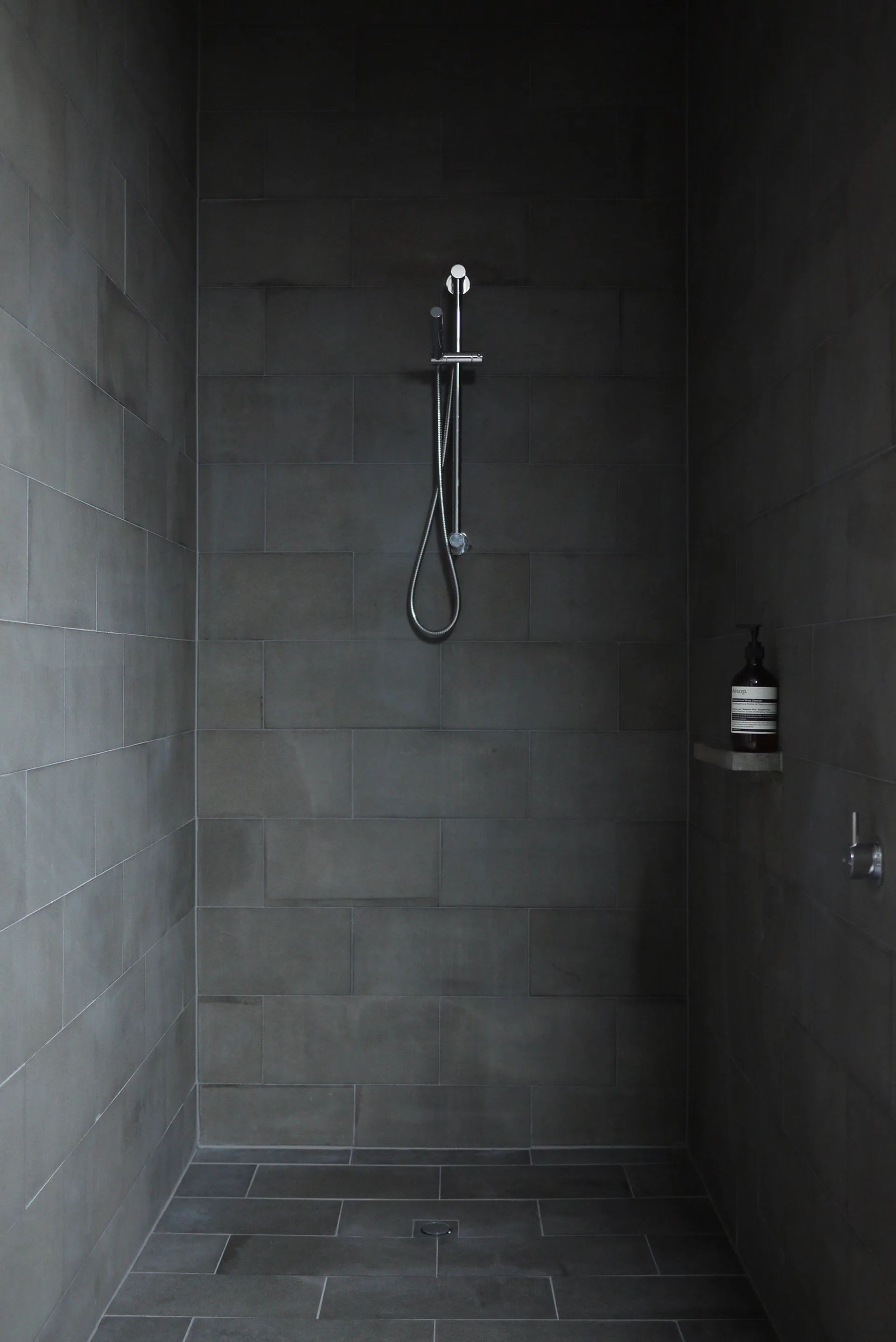 A minimalist shower stall features matte charcoal grey tiles on the walls and floor with a silver shower head and faucet.