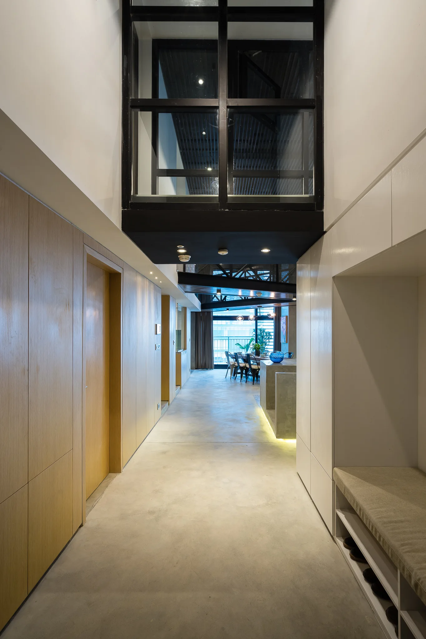 A modern hallway with light wood paneling and a white built-in bench leads to a dining area over a smooth grey concrete floor.