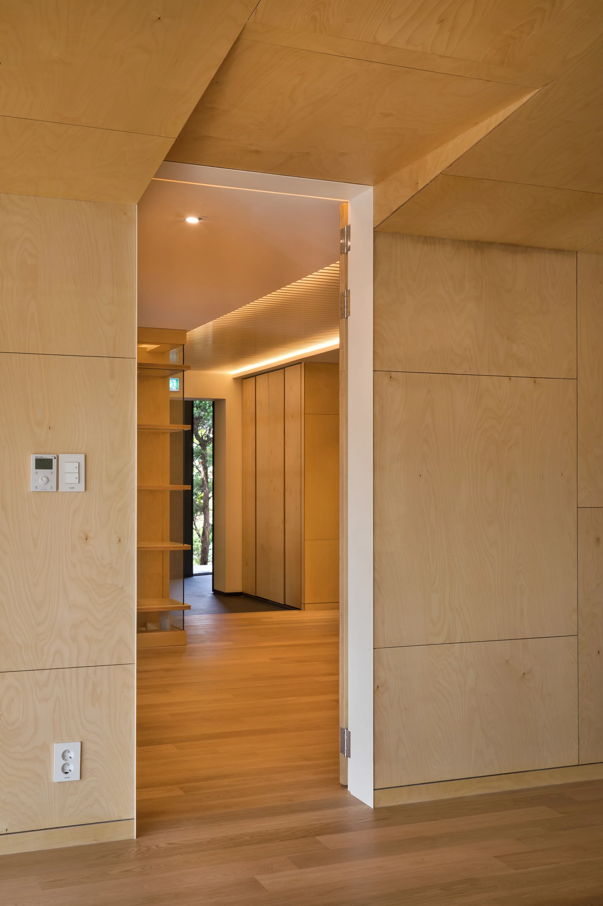 A modern doorway looks into a hallway featuring light-toned plywood wall panels, matching wood floors, and integrated ceiling cove lighting.