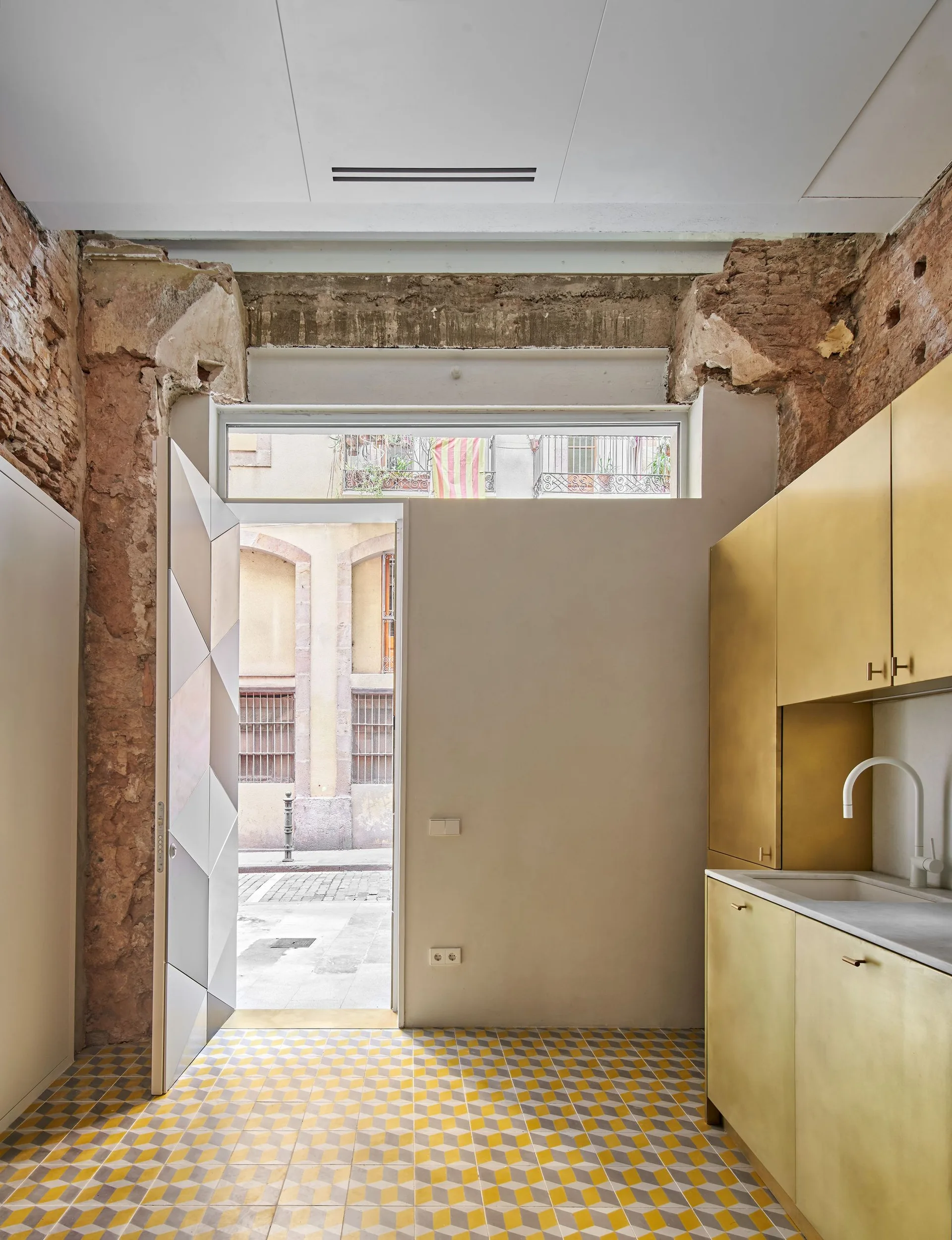 A modern kitchen interior features gold-toned metallic cabinetry, a geometric cube-patterned floor, and raw exposed brick surrounding an open doorway.