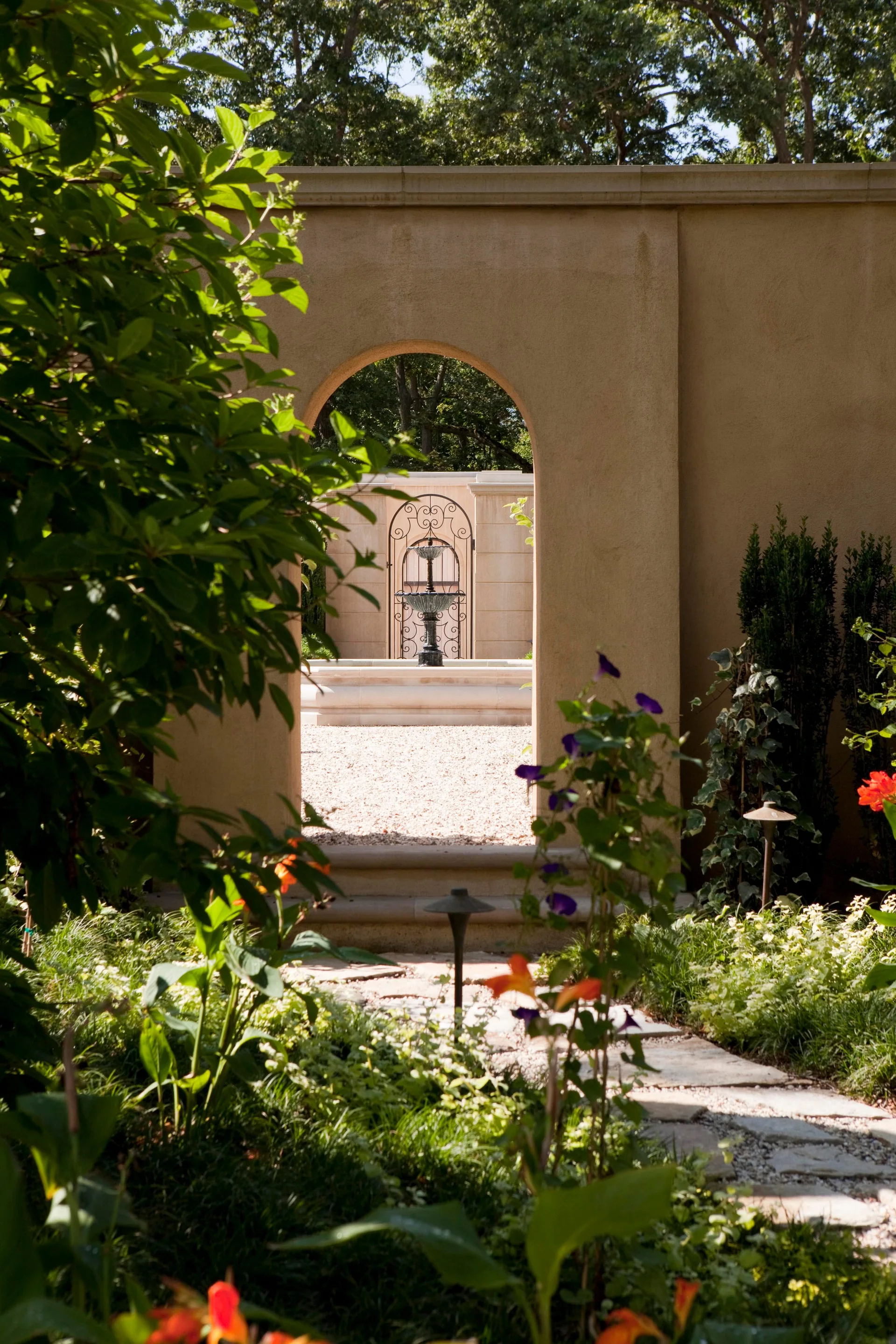 An arched opening in a tan stucco wall looks onto a gravel courtyard featuring a tiered black fountain in front of an iron gate.