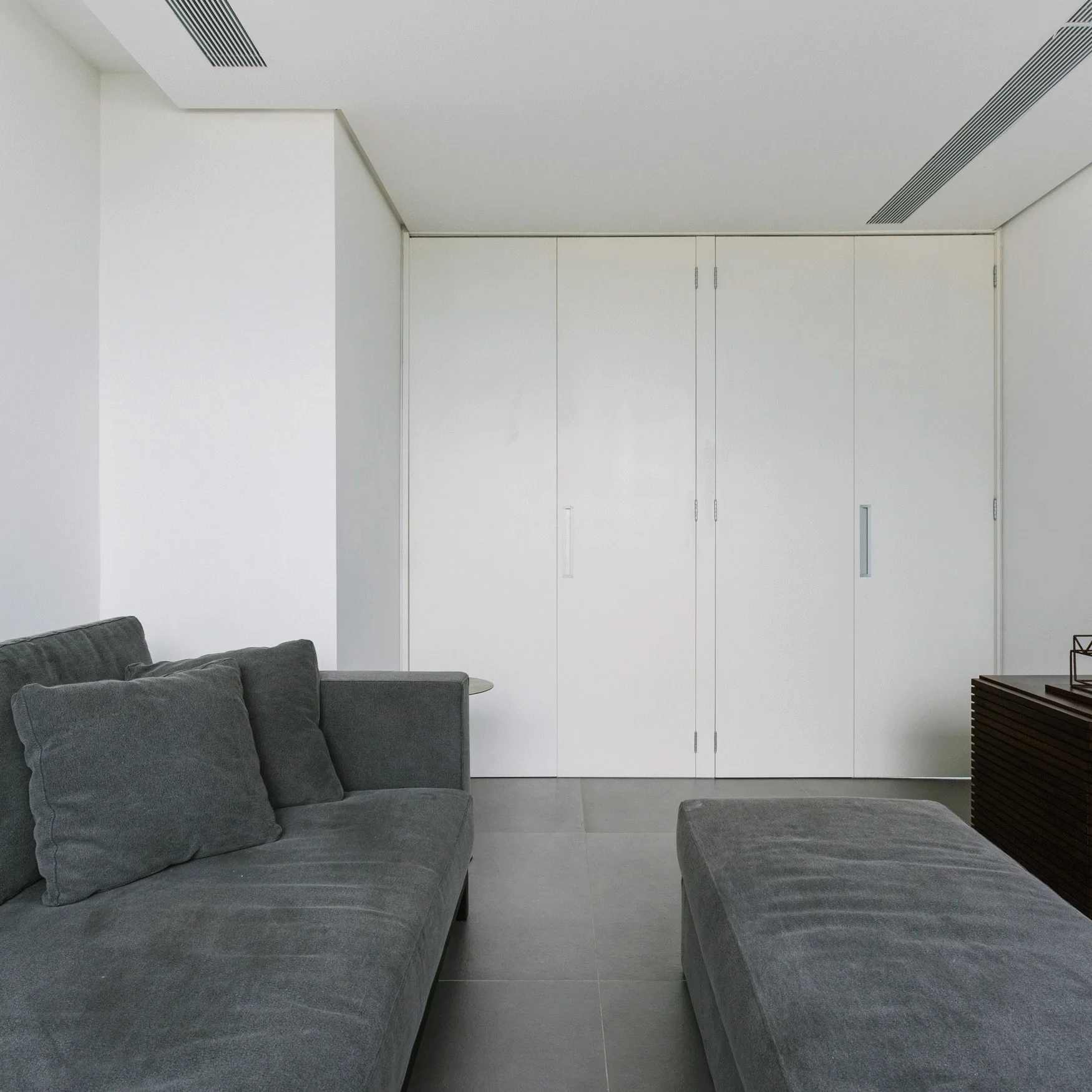 A minimalist living room features a grey fabric sofa and ottoman facing a wall of floor-to-ceiling white cabinets with simple silver handles.
