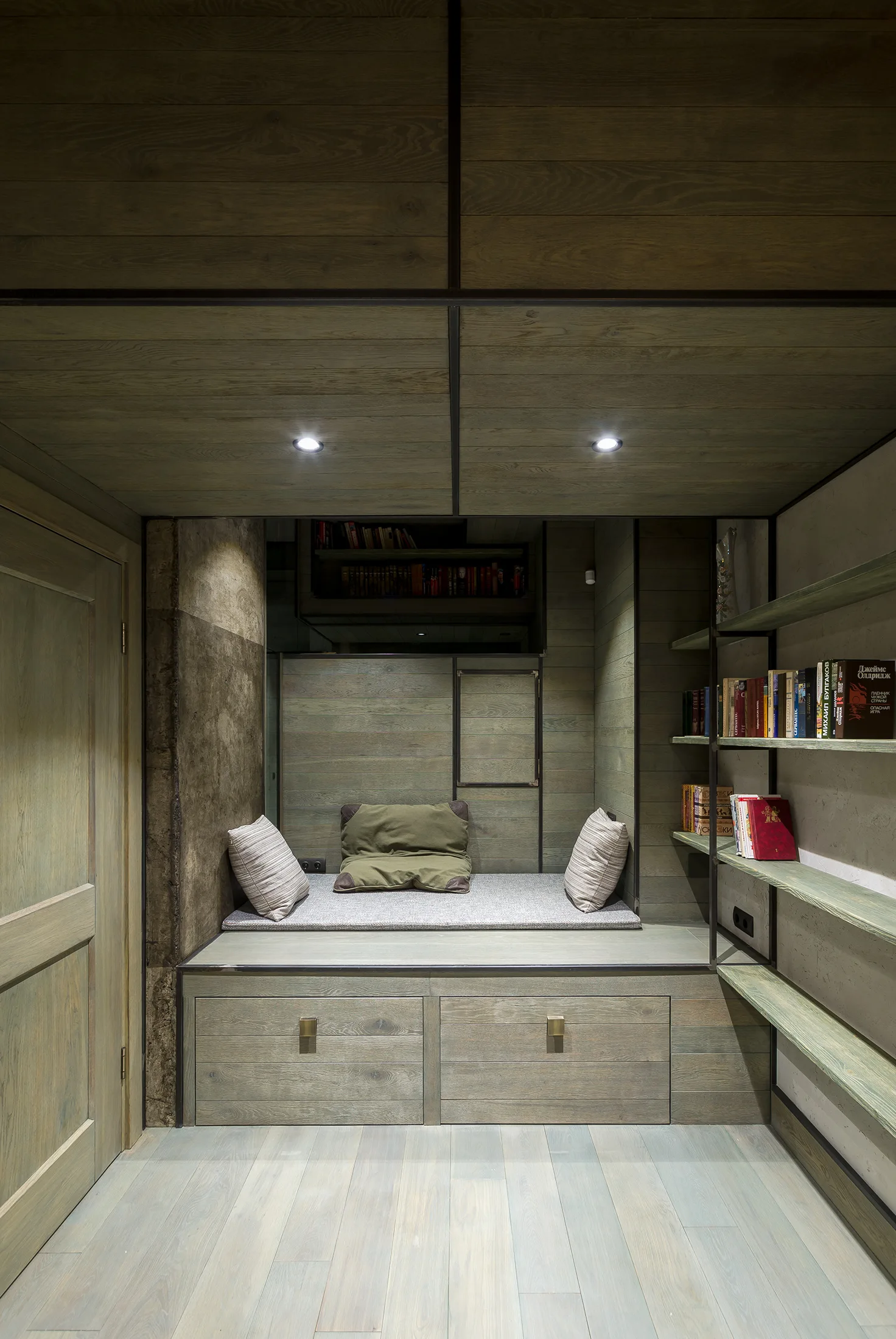 A cozy reading nook features a built-in daybed with large drawers below and integrated wooden shelves against a muted green plank wall.