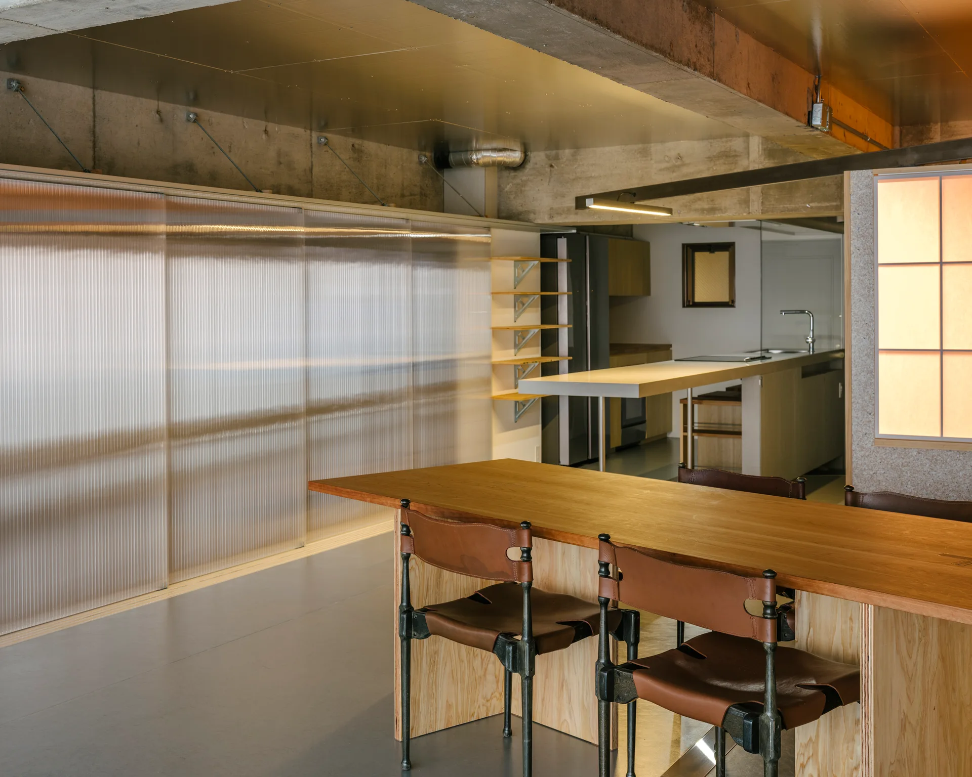 An industrial dining area with a long wooden table, leather-strapped chairs, and a large corrugated translucent sliding partition.