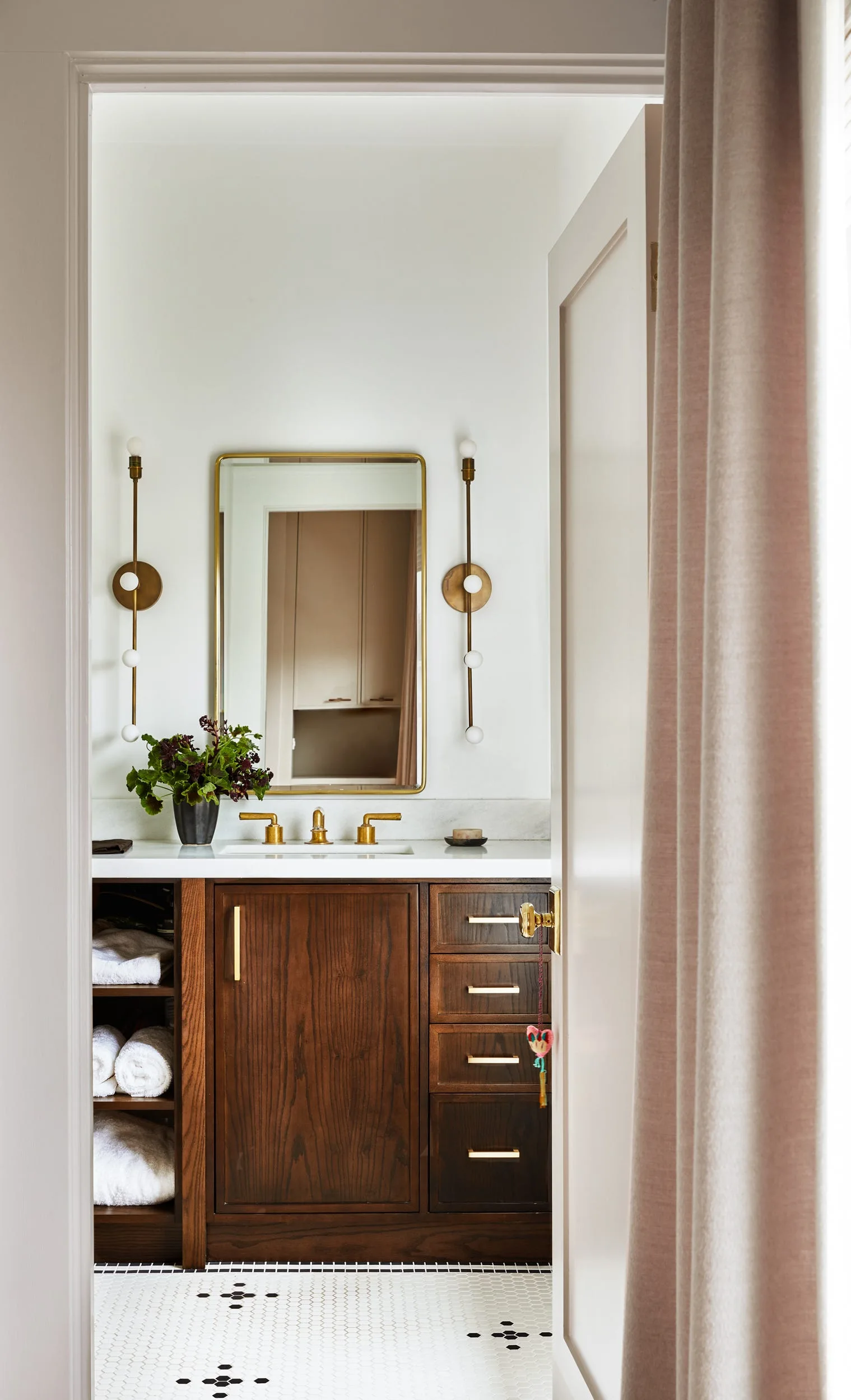 A dark wood bathroom vanity features brass faucets, a brass-framed mirror, and vertical wall sconces above white hexagonal floor tiles.