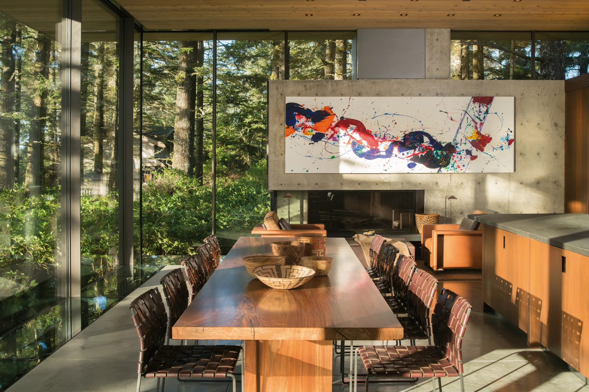 A long wood slab dining table with woven leather chairs sits in an open-concept room featuring floor-to-ceiling glass walls looking onto a pine forest.
