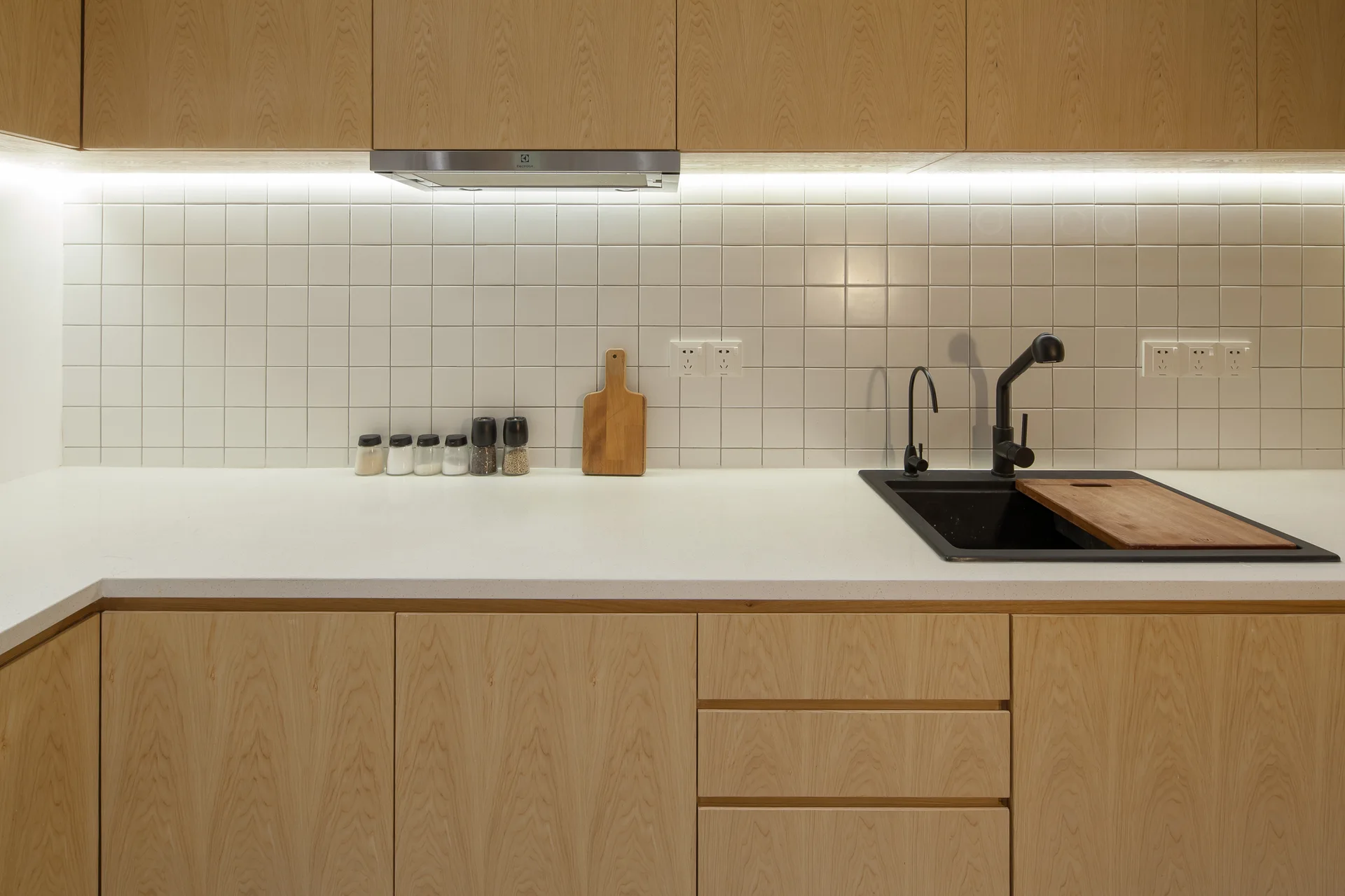 A modern kitchen features light wood cabinets, a white countertop, a white square-tile backsplash, and a black sink with a matching black faucet.