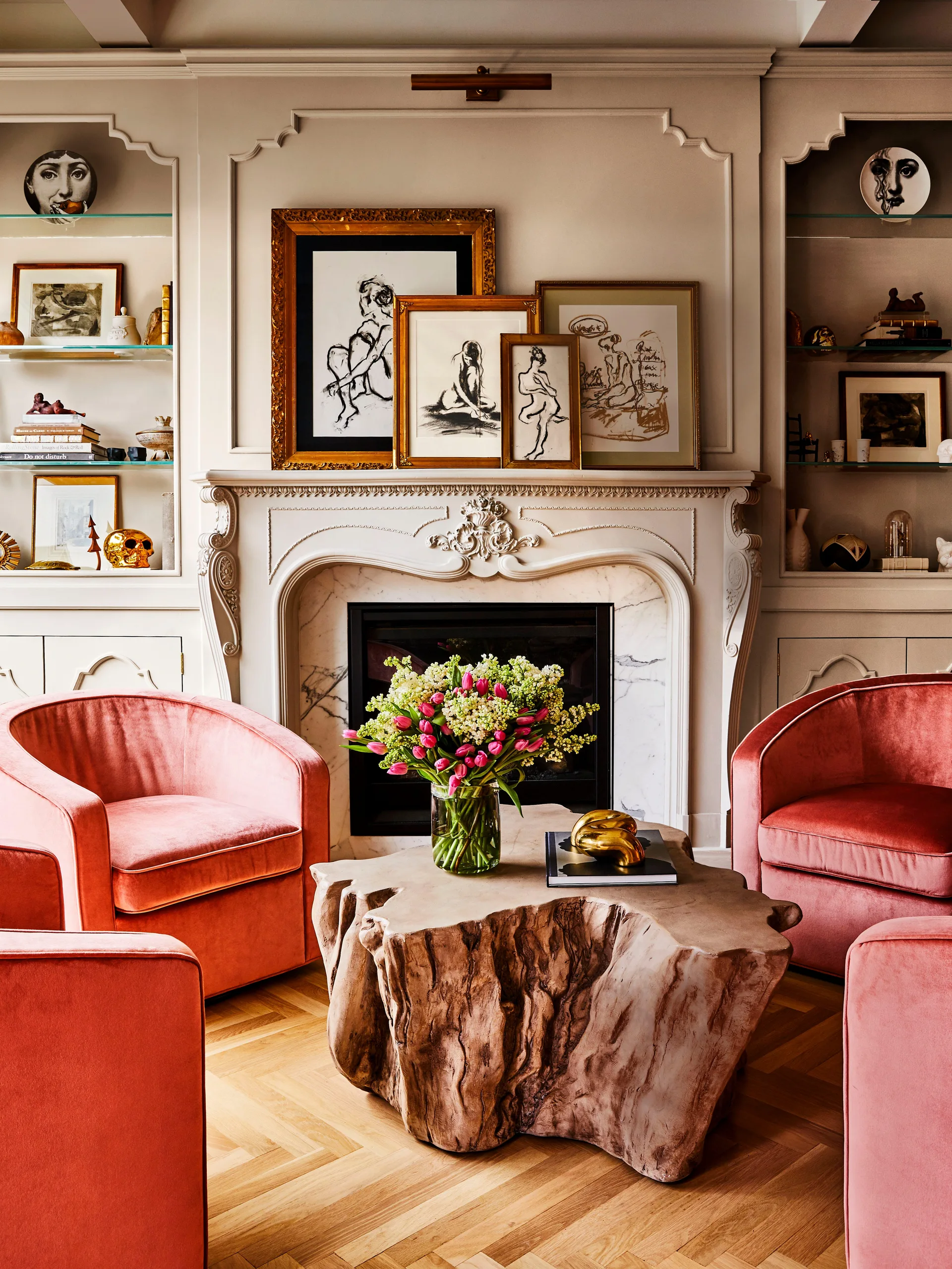 A classical fireplace with symmetrical built-in shelving is paired with pink velvet armchairs and an organic wood coffee table.