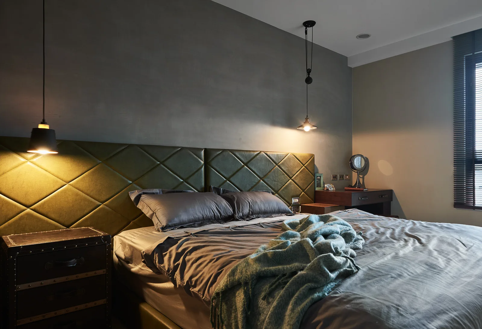 A moody bedroom featuring an olive green tufted headboard against a gray wall, styled with warm pendant lights and a trunk-style nightstand.