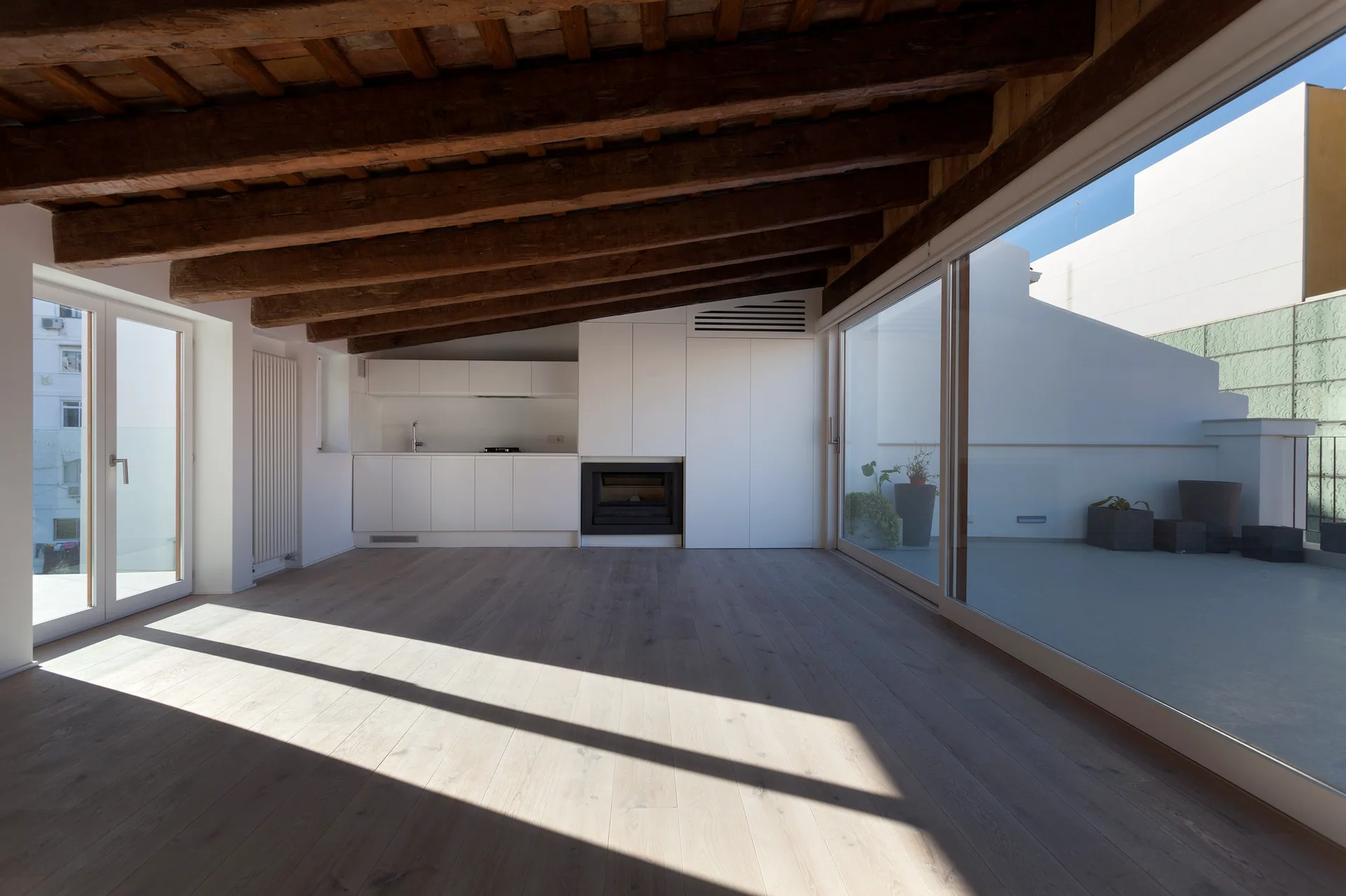 A bright minimalist living space with exposed dark wood ceiling beams, light wood floors, and large sliding glass doors leading to an outdoor terrace.
