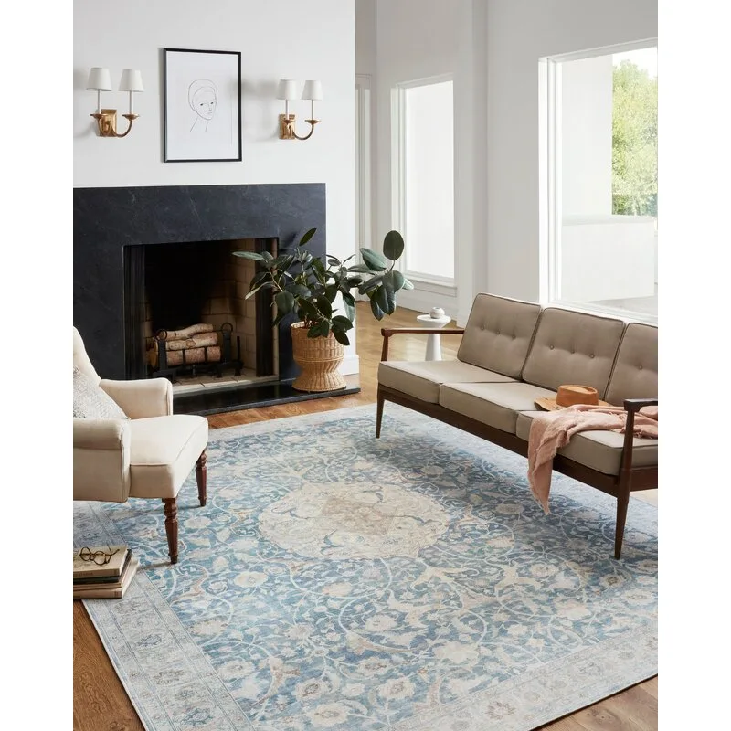 A bright living room features a blue patterned area rug, a tufted mid-century modern sofa, an armchair, and a dark stone fireplace.