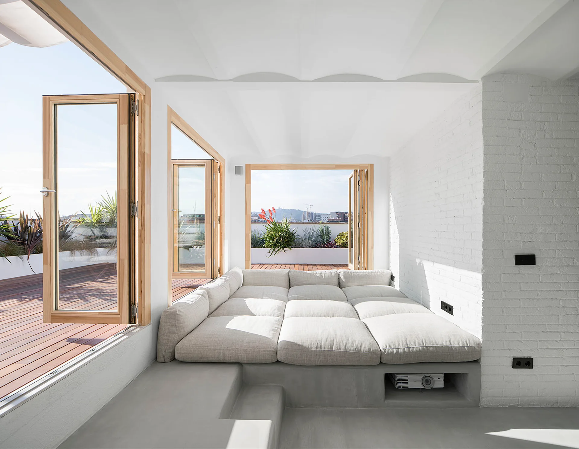A modular grey daybed on a raised concrete platform next to wood-framed folding glass doors that open to a sunny rooftop terrace.