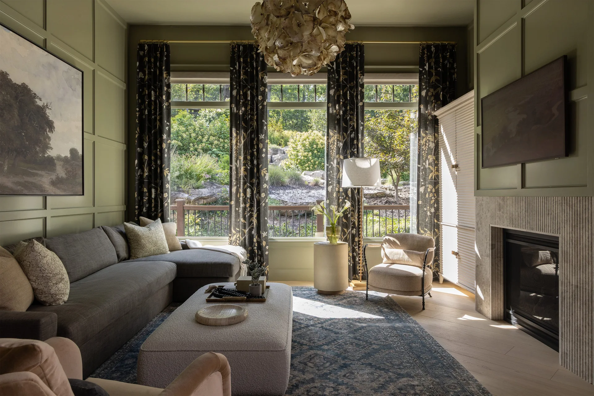 A sage green living room features a grey sectional sofa, an organic shell chandelier, botanical print curtains, and a fluted stone fireplace.