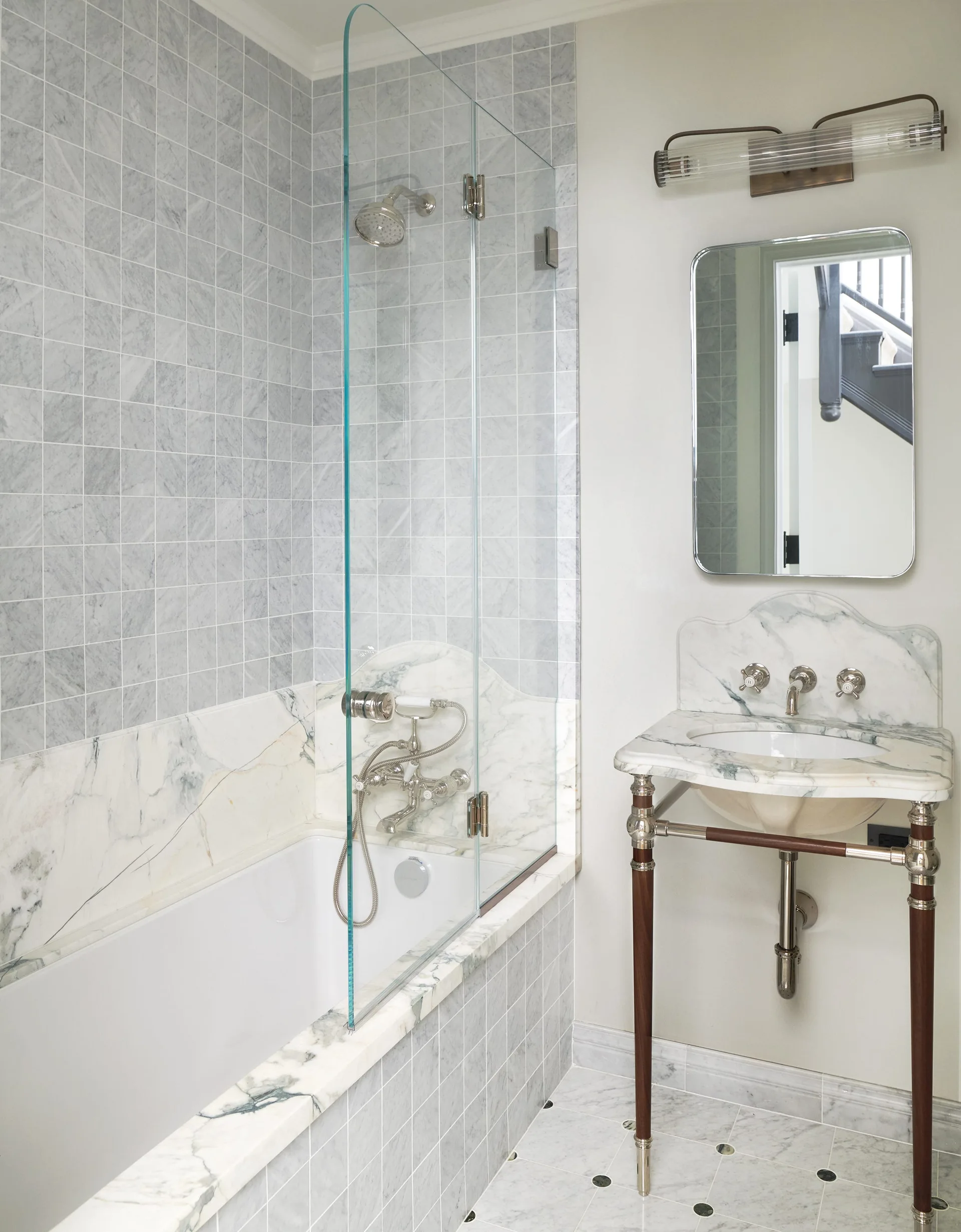 A light grey marble bathroom features a console sink with dark wood legs next to a bathtub with a clear glass shower screen.