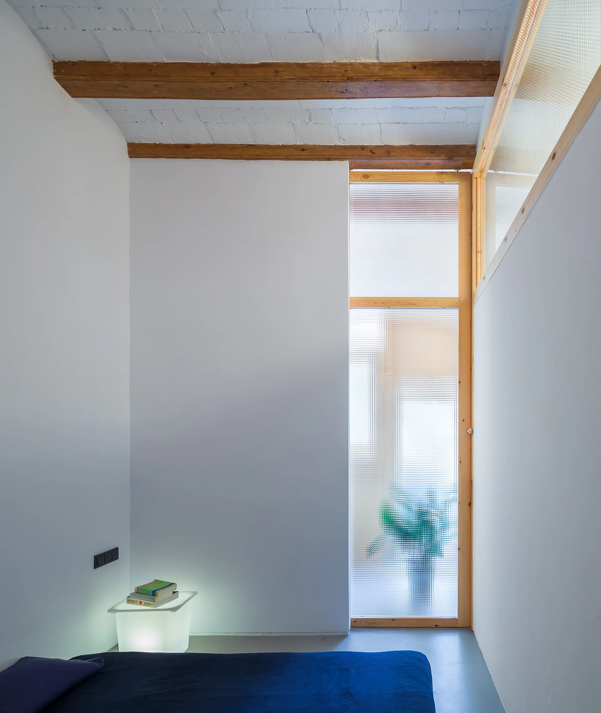 A minimalist bedroom featuring a blue bed, a glowing cube nightstand, and a textured glass door under exposed wooden ceiling beams.