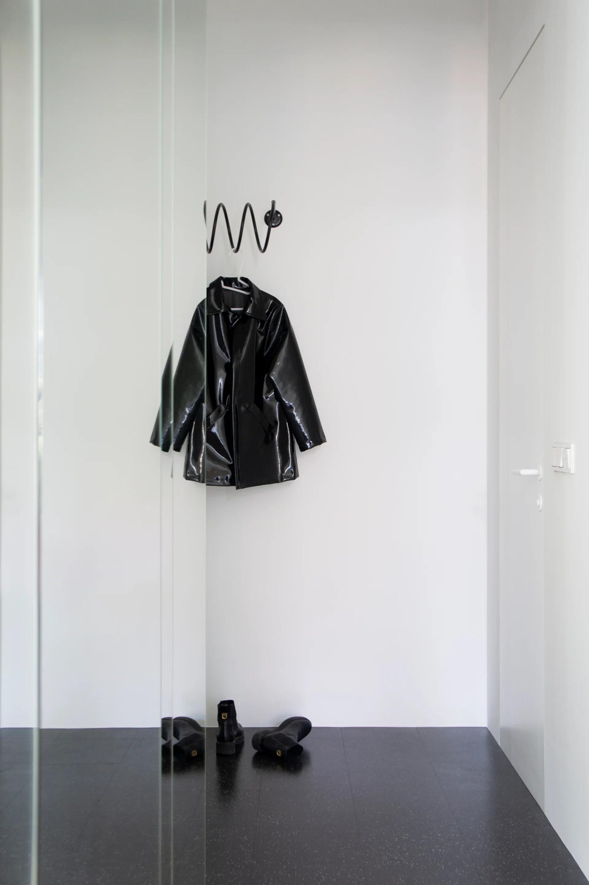 A black glossy coat hangs from a spiral metal wall hook in a minimalist white entryway with dark tile flooring and a pair of black boots.