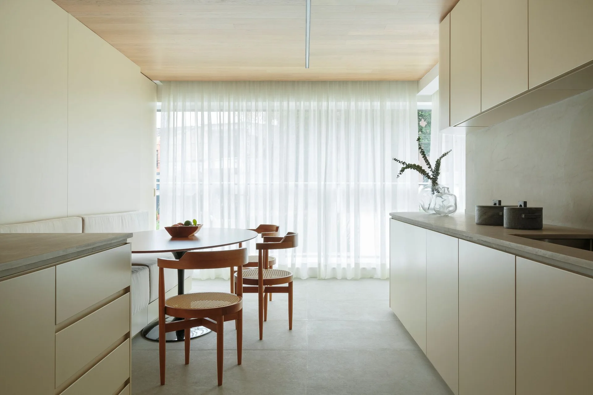 A modern dining area features a built-in bench, a round wooden table, cane chairs, and a light wood paneled ceiling next to sheer white curtains.