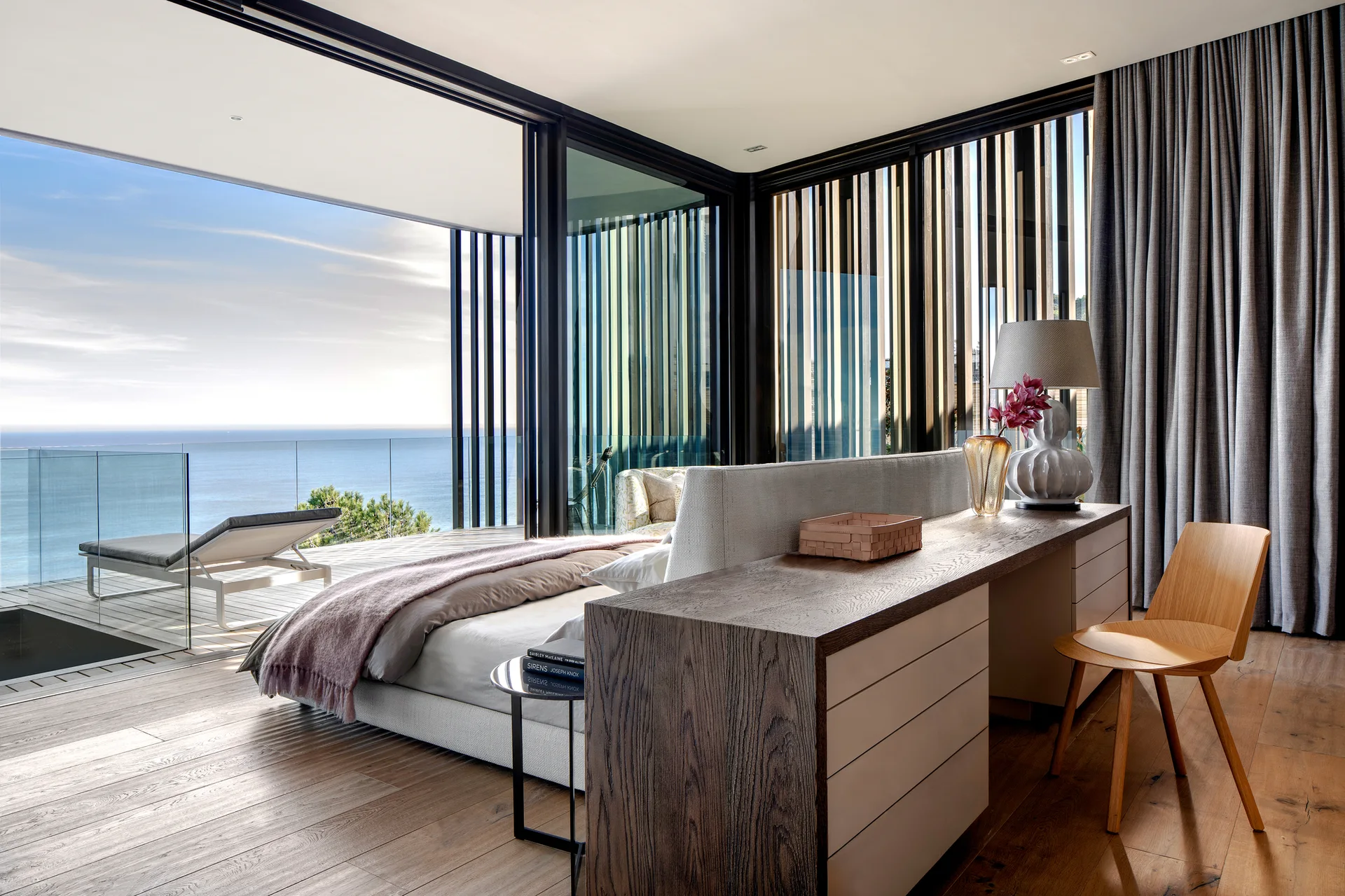 A modern bedroom features an upholstered bed with a dark wood desk positioned behind the headboard, opening onto a terrace with ocean views.