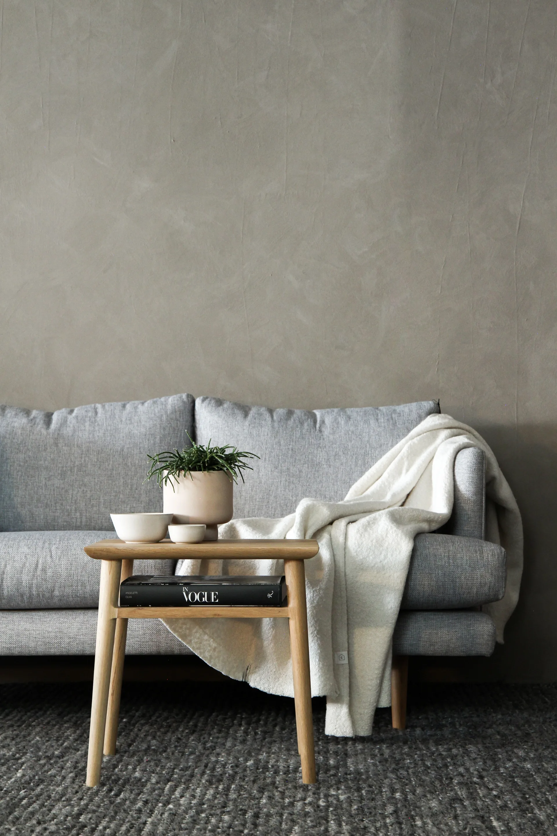A gray fabric sofa stands against a textured gray plaster wall next to a wooden side table decorated with a potted plant and ceramic bowls.