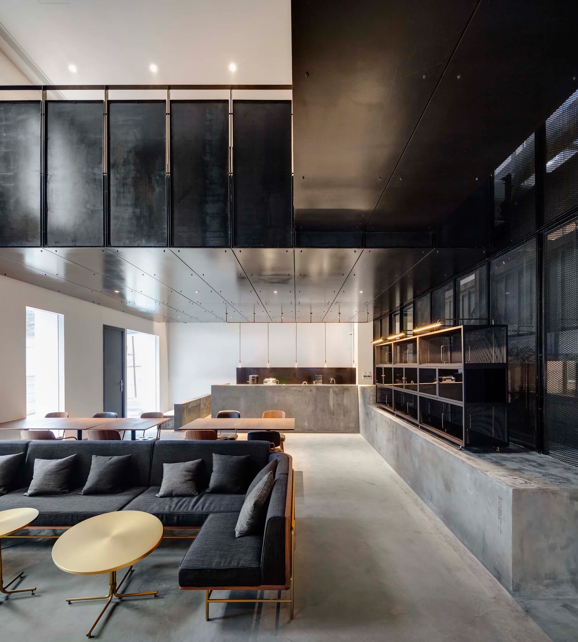 A modern industrial living space features a charcoal sectional sofa with round brass coffee tables on a concrete floor beneath dark metal ceiling panels.
