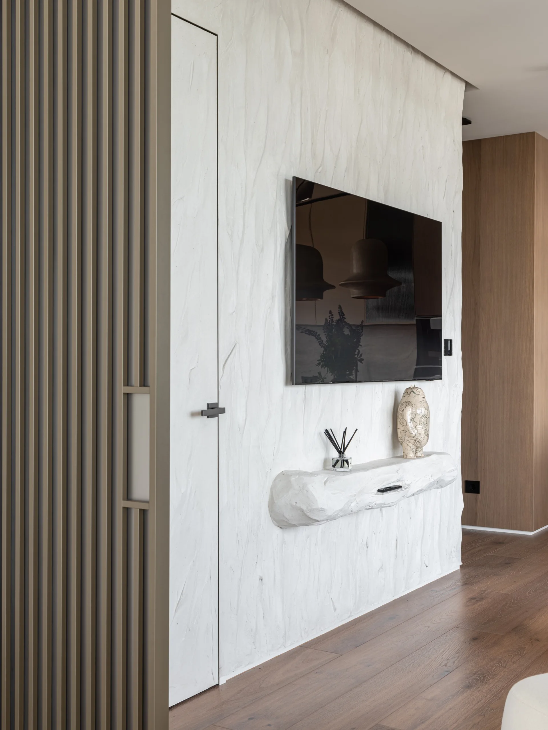 A white textured plaster feature wall with an integrated flush door, a wall-mounted flat-screen television, and an organic-shaped floating shelf.