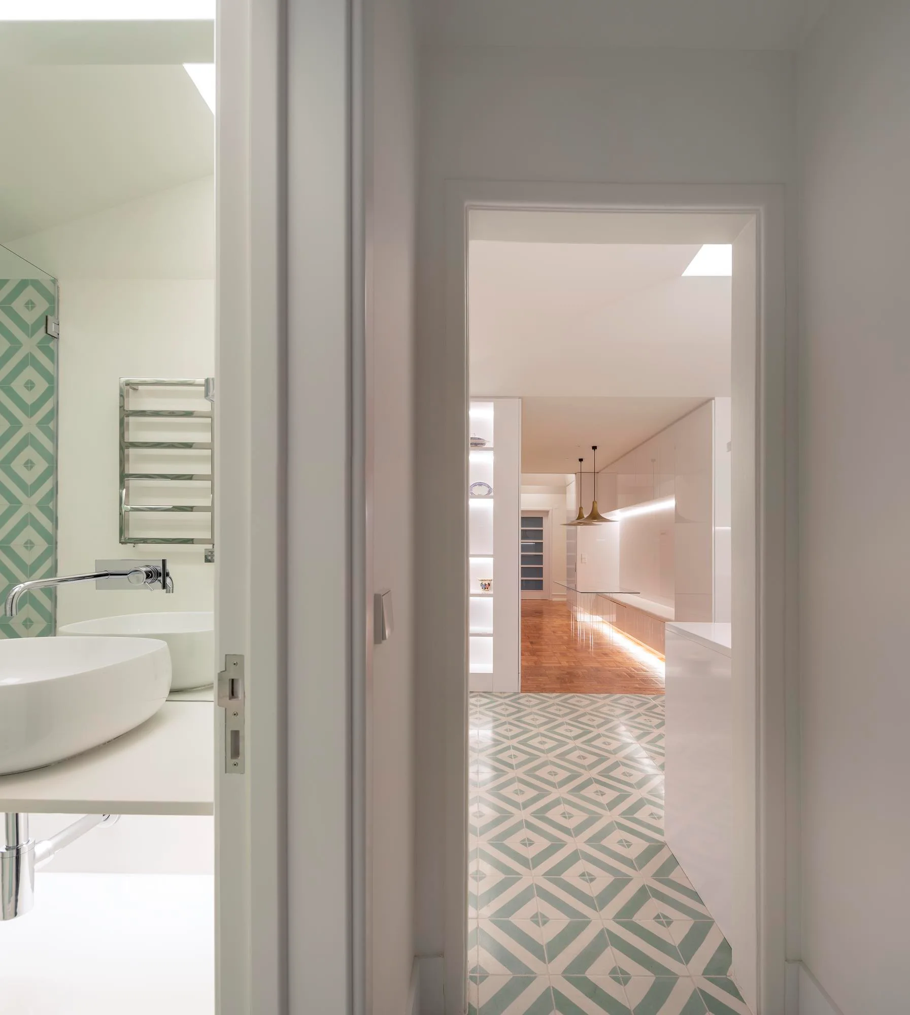 A view through a white doorway showing a bathroom with a white sink and green geometric floor tiles leading into a hallway.