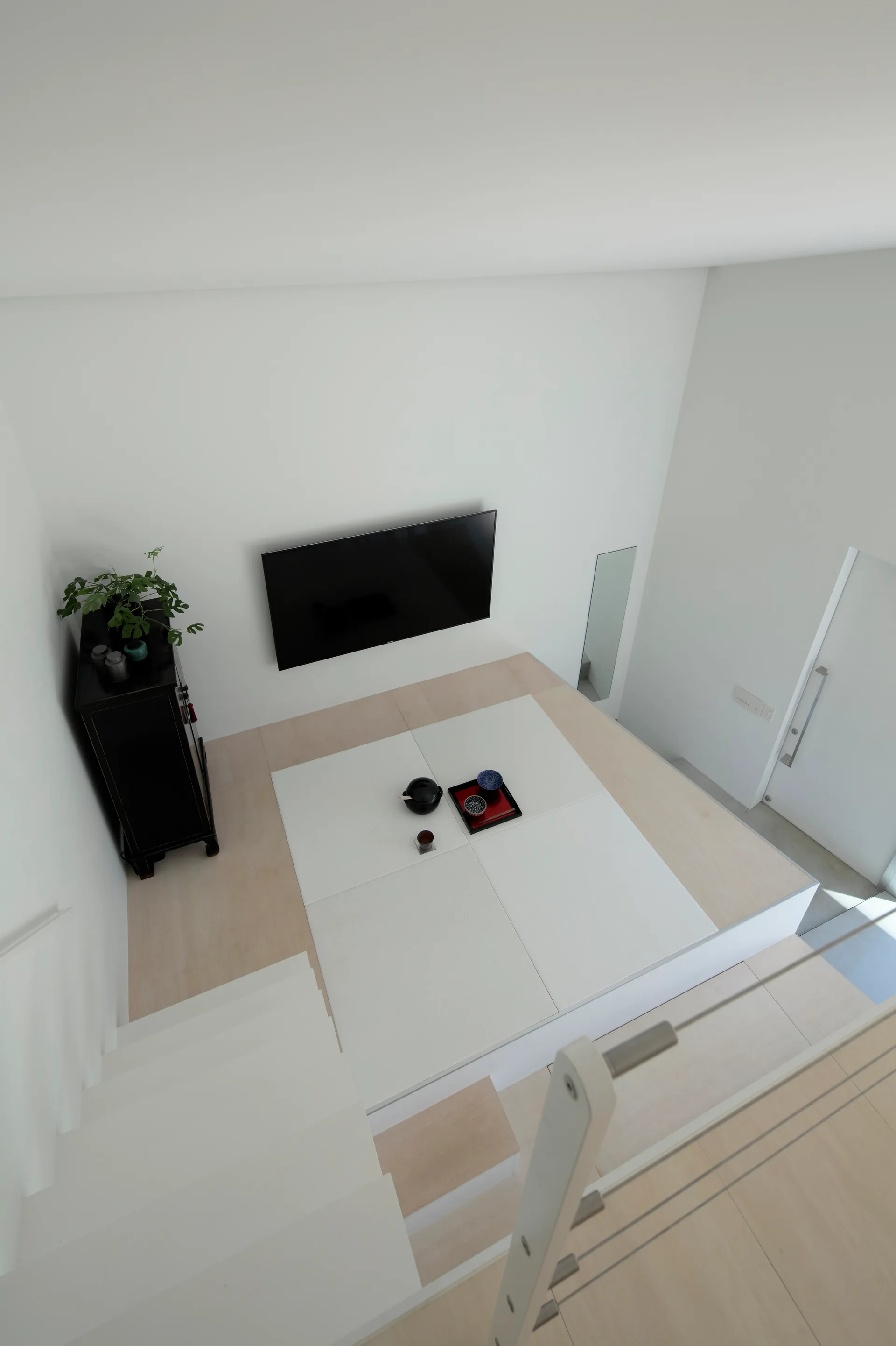 A high-angle shot of a minimalist white room featuring an elevated light wood platform with inset white tatami mats and a wall-mounted TV.