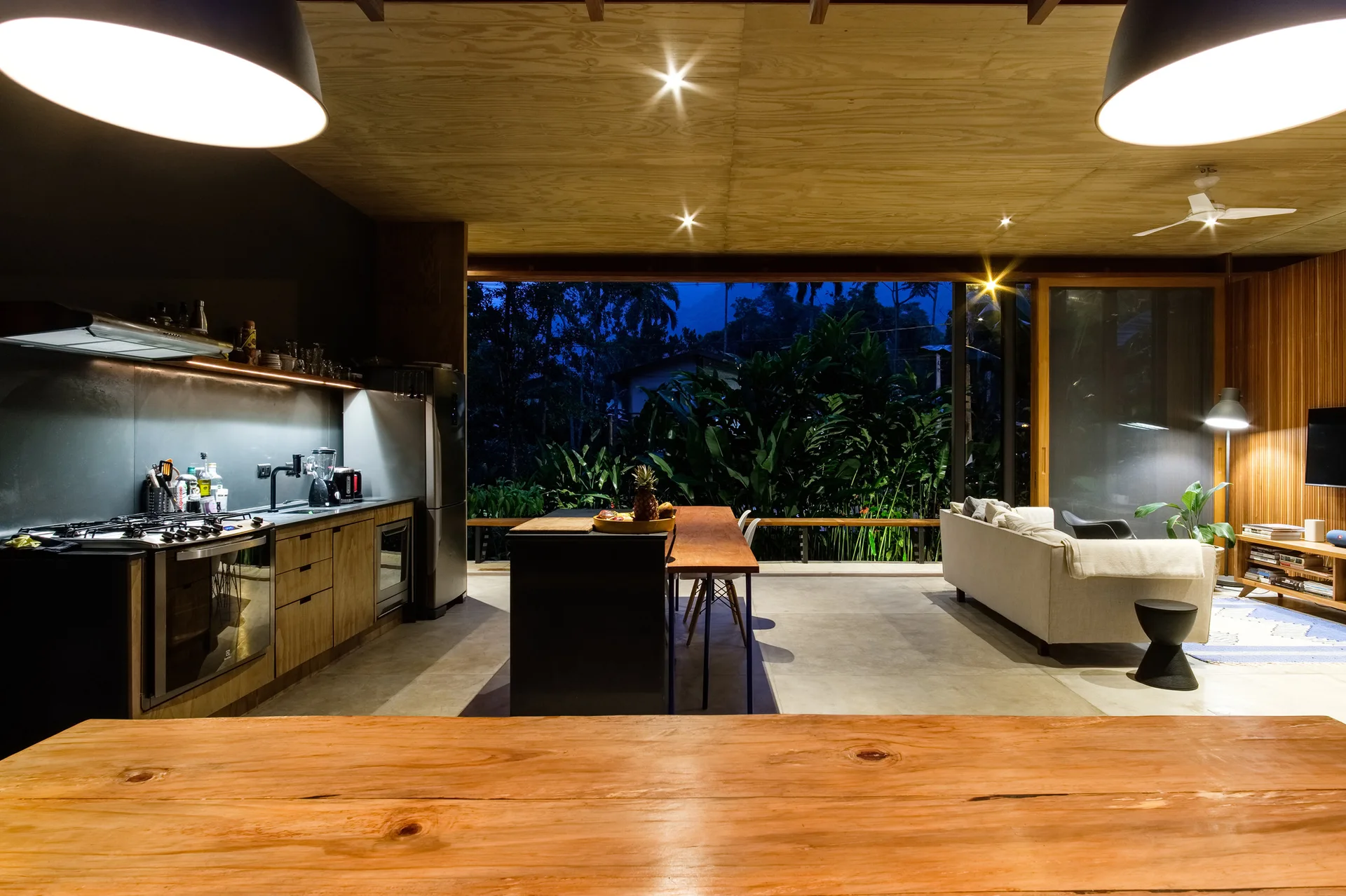 An open plan kitchen with a wood plank ceiling, a black island, and large glass walls overlooking a tropical landscape at night.