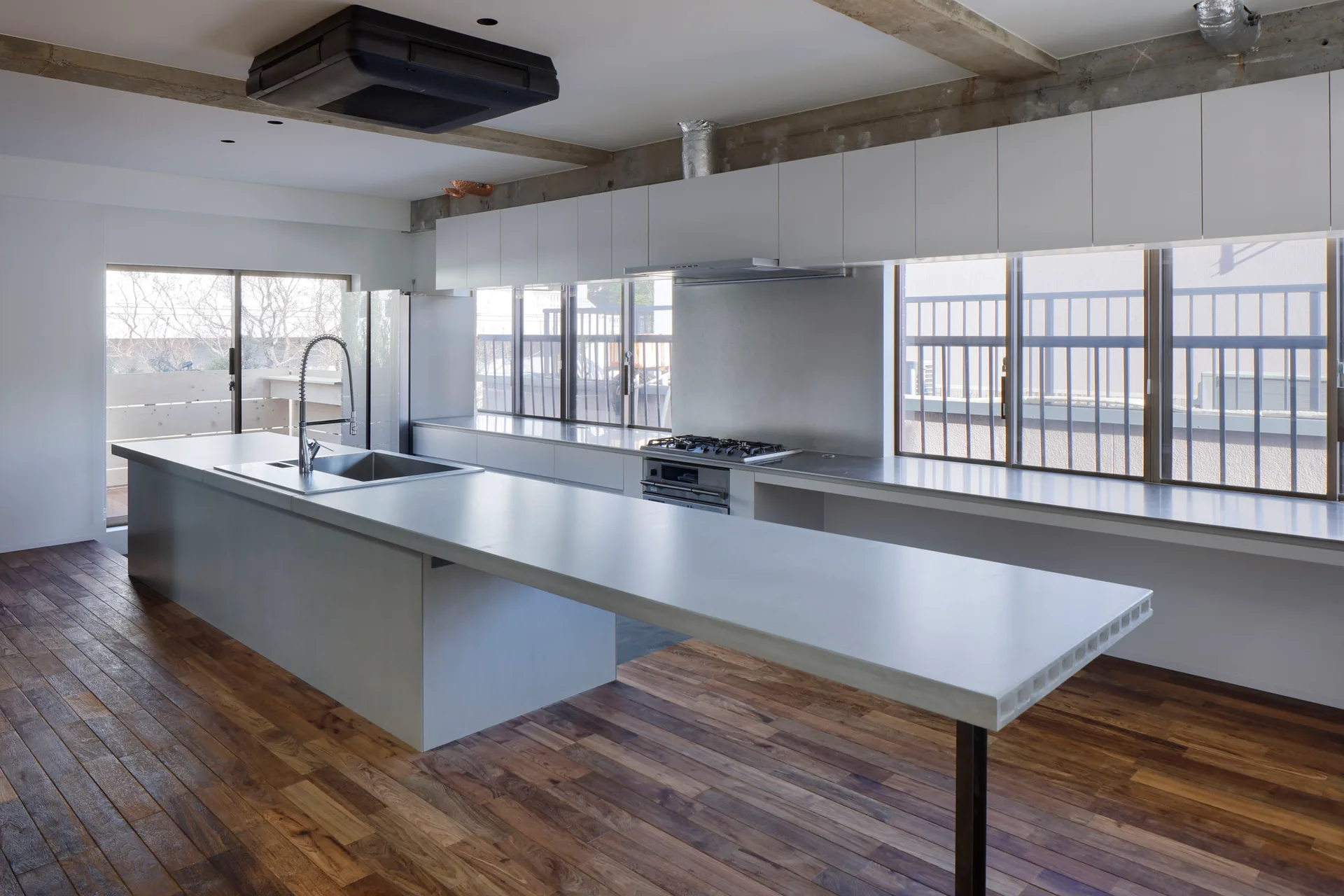 A minimalist kitchen featuring a long grey concrete-look island, white cabinetry, exposed concrete ceiling beams, and warm wood flooring.