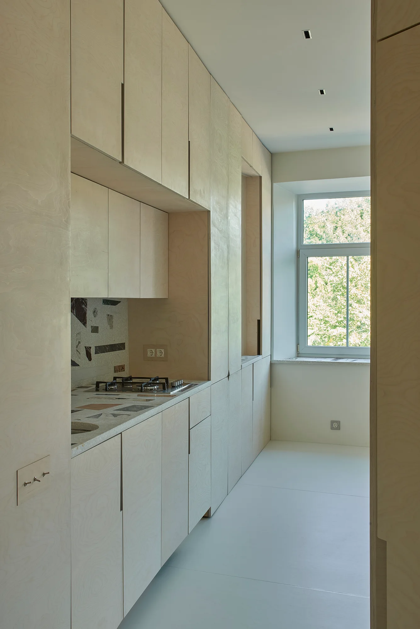 A minimalist galley kitchen featuring light birch plywood cabinetry, an integrated gas cooktop with a terrazzo backsplash, and a window at the far end.