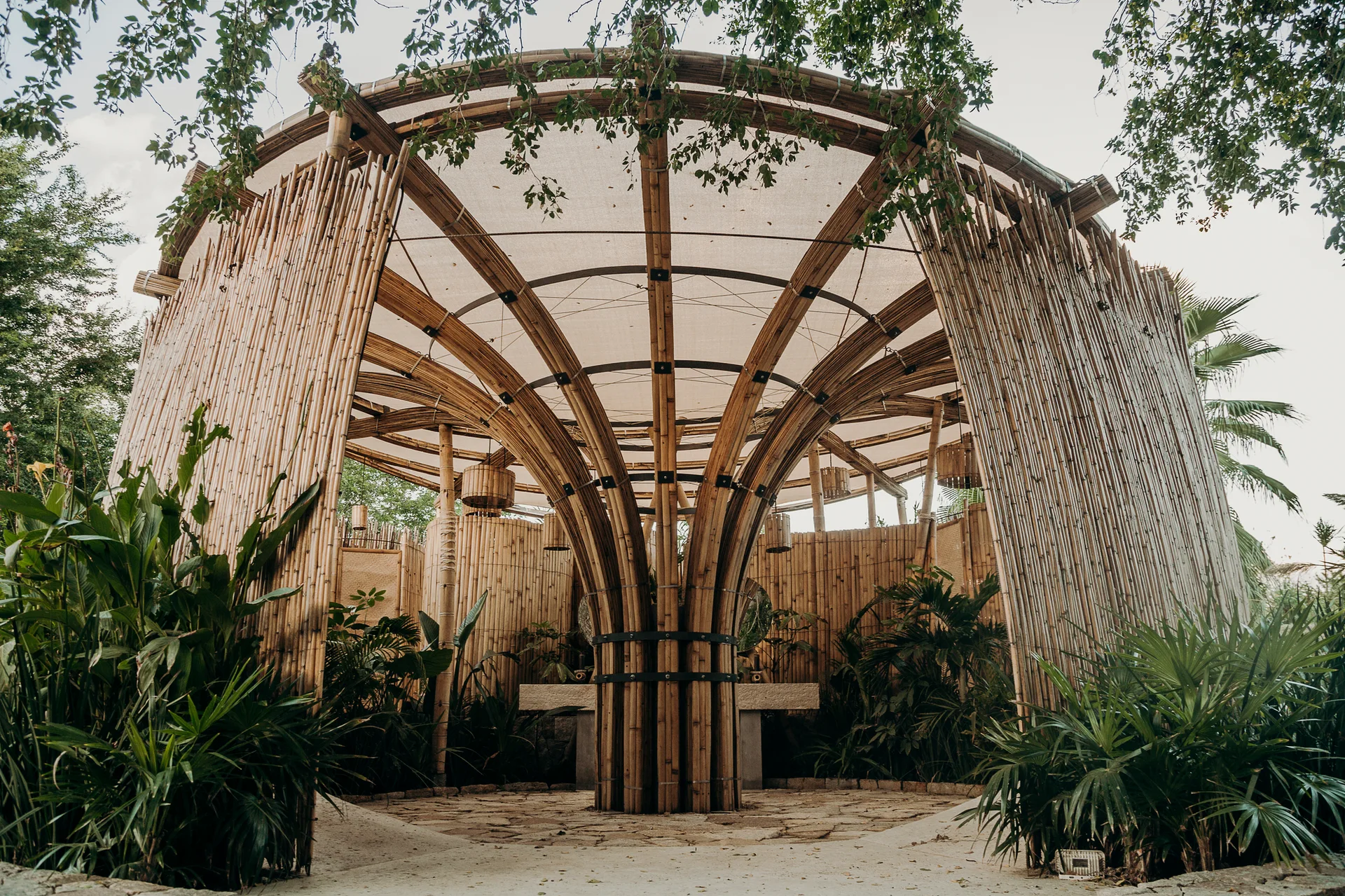 An open-air circular bamboo pavilion features a central tree-like column supporting a fabric canopy roof surrounded by tropical plants.