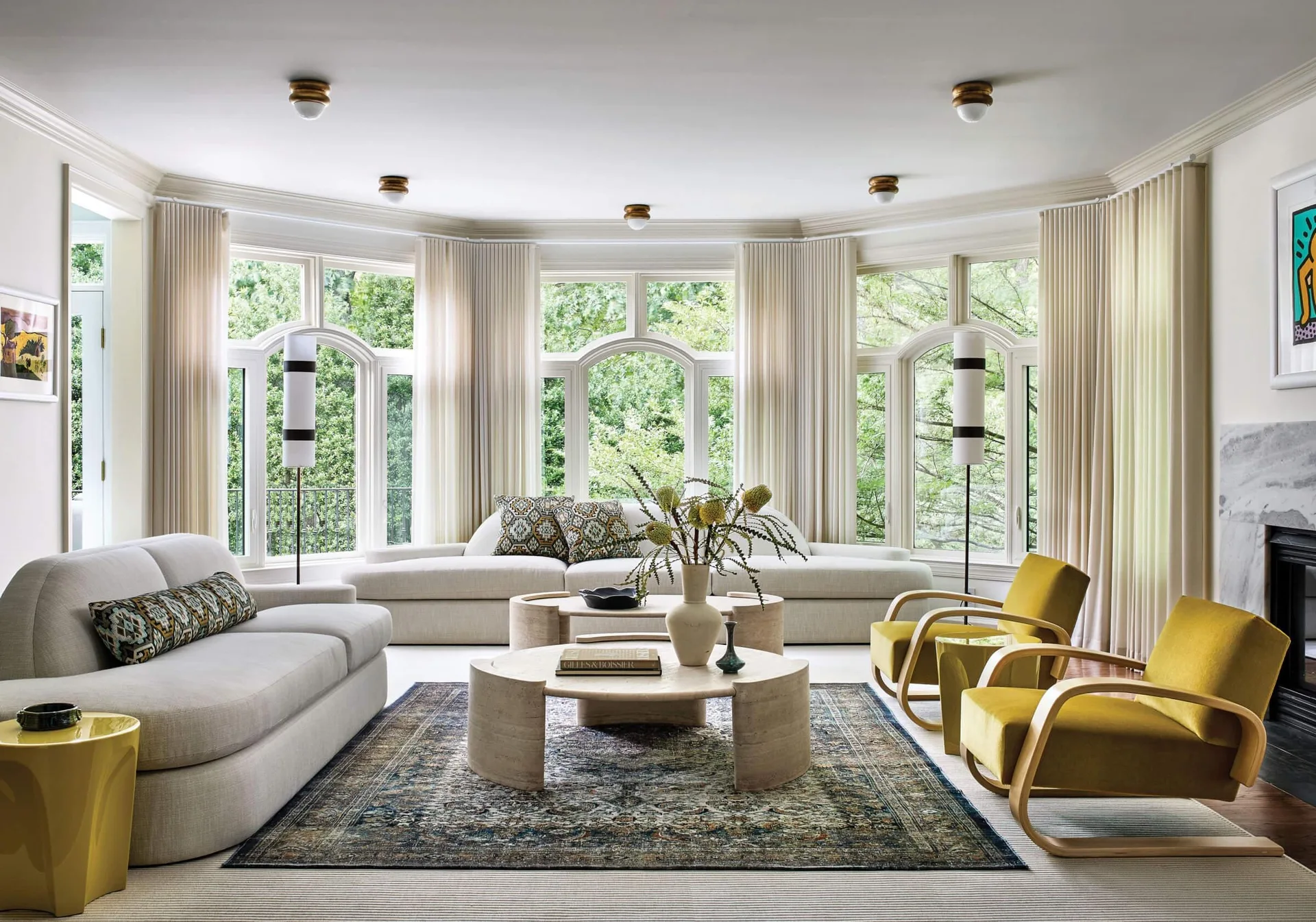 A bright living room features cream sofas, mustard yellow velvet armchairs, and round travertine coffee tables in front of a large curved bay window.