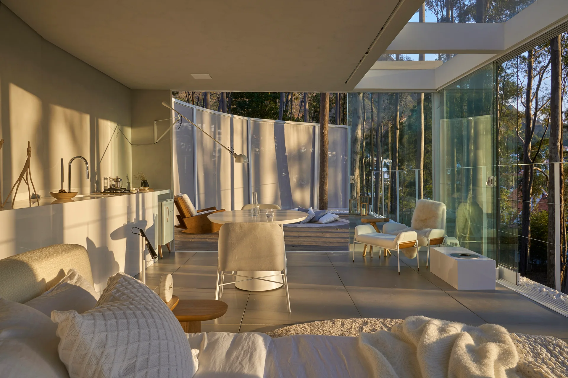 A modern bedroom with floor-to-ceiling glass doors opening onto a wooden terrace with a forest view, featuring a bed, small dining table, and armchair.