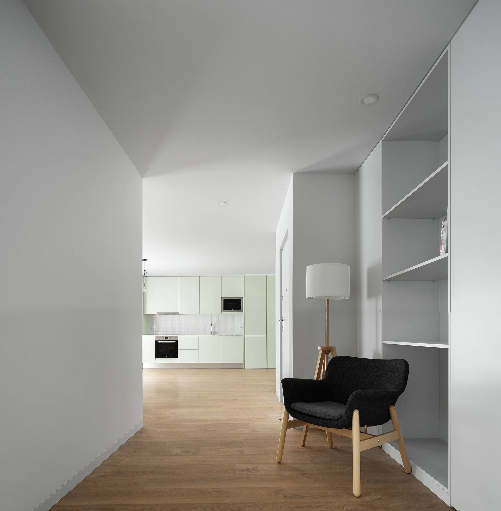 A minimalist hallway features a dark gray armchair and tripod lamp next to white shelving, with a pale green kitchen in the background.