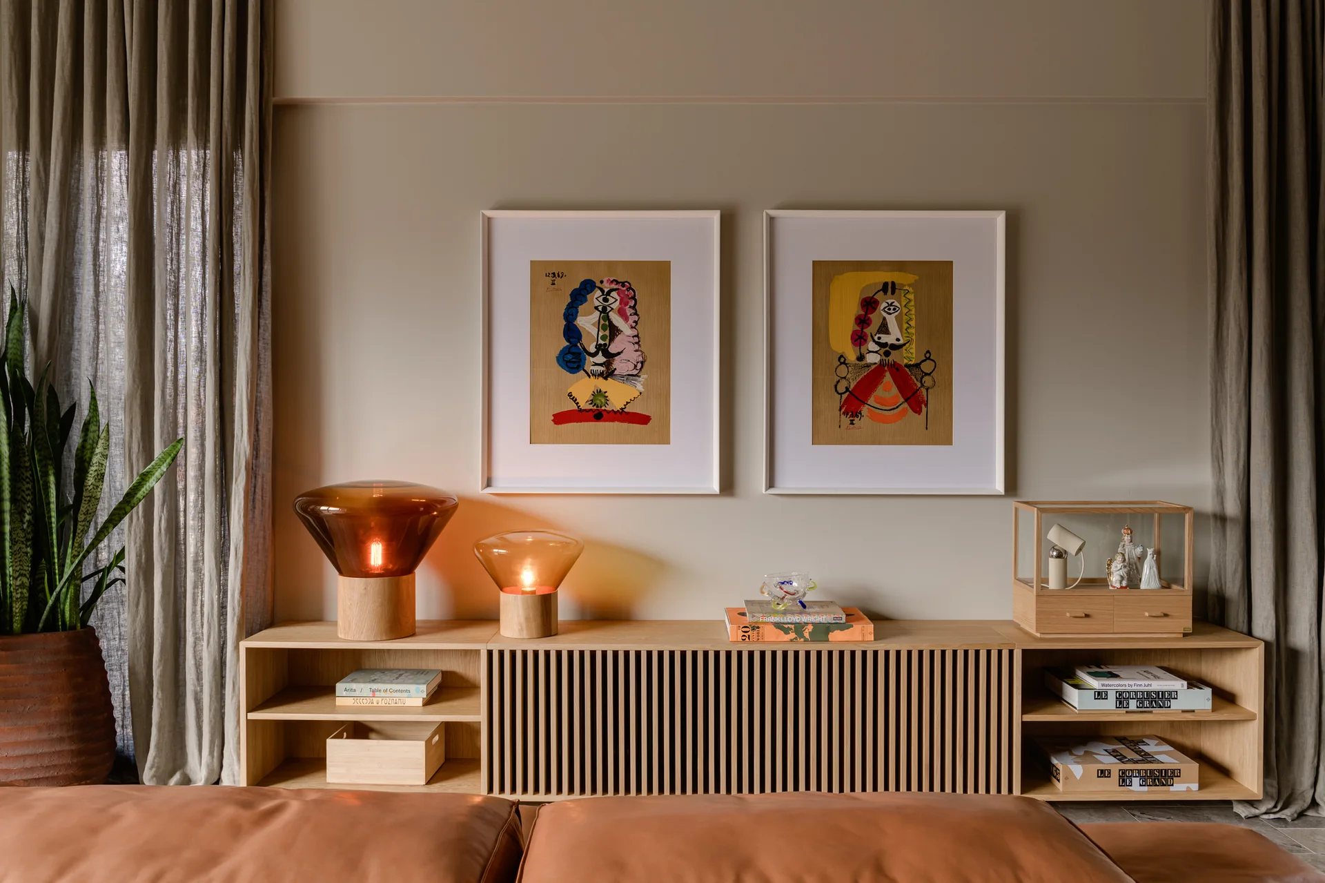 A low oak console table with vertical slats is styled with two glowing amber glass table lamps and framed colorful art on a beige wall.