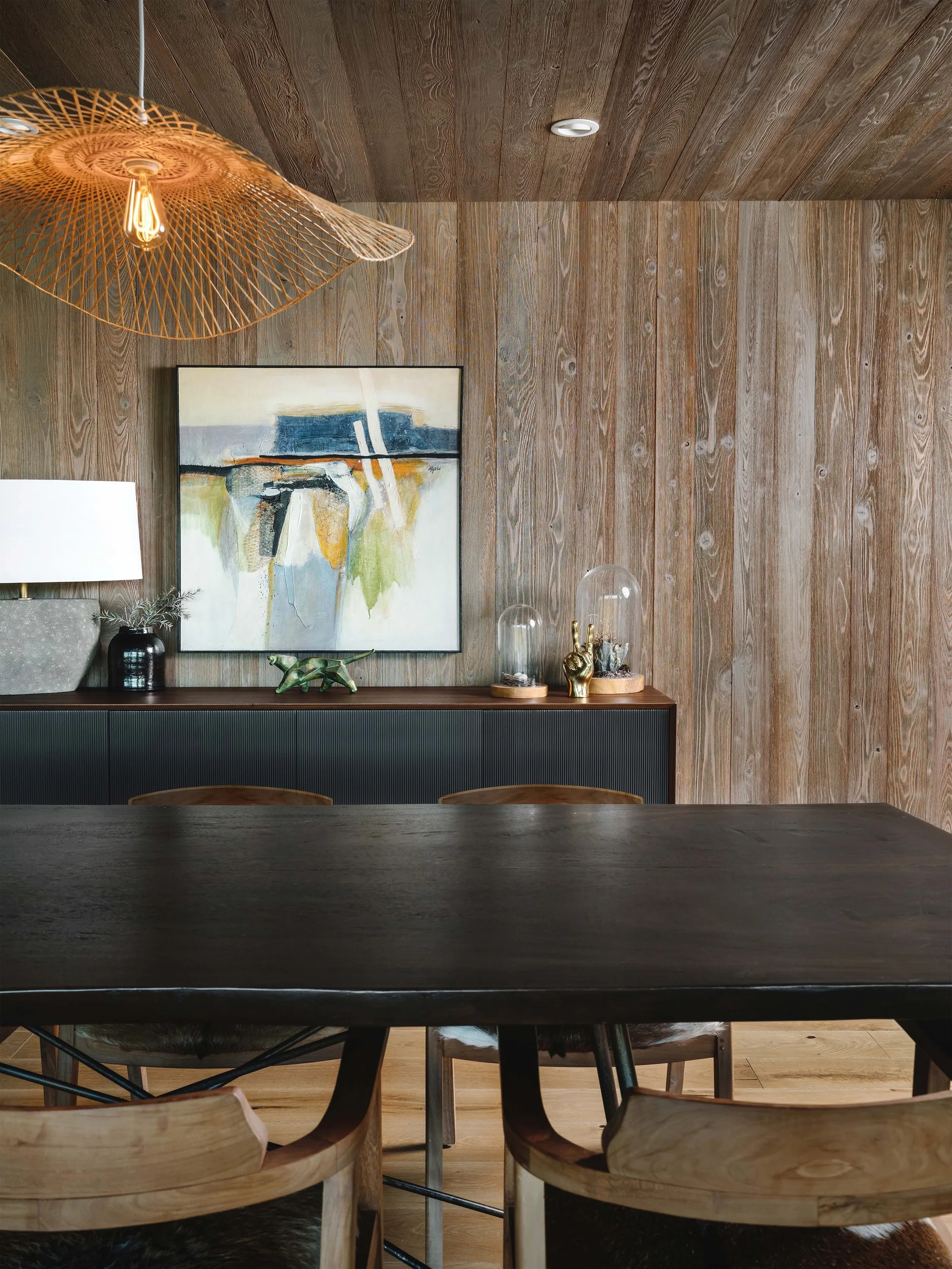 A dining room features vertical wood planking on the walls and ceiling, a dark dining table, and a ribbed black sideboard beneath abstract art.