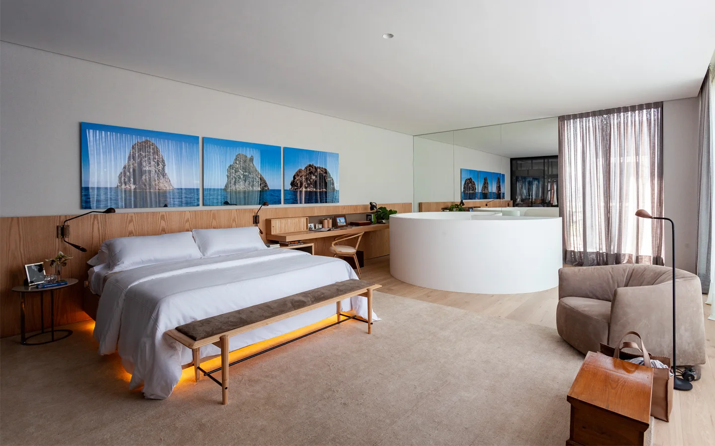 A modern bedroom features a bed with wood wall paneling, a floating desk, and a white circular bathtub enclosed by a curved partition.