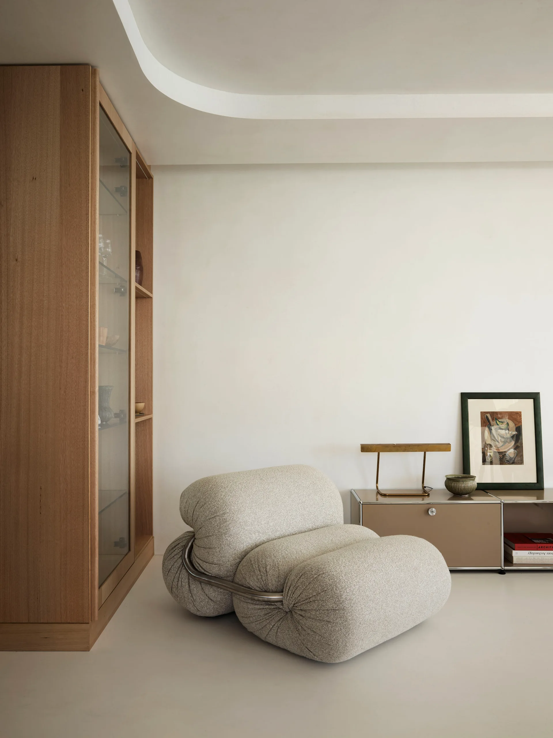 A plush textured grey armchair sits beside a modular beige sideboard and a tall oak display cabinet against plain white walls.