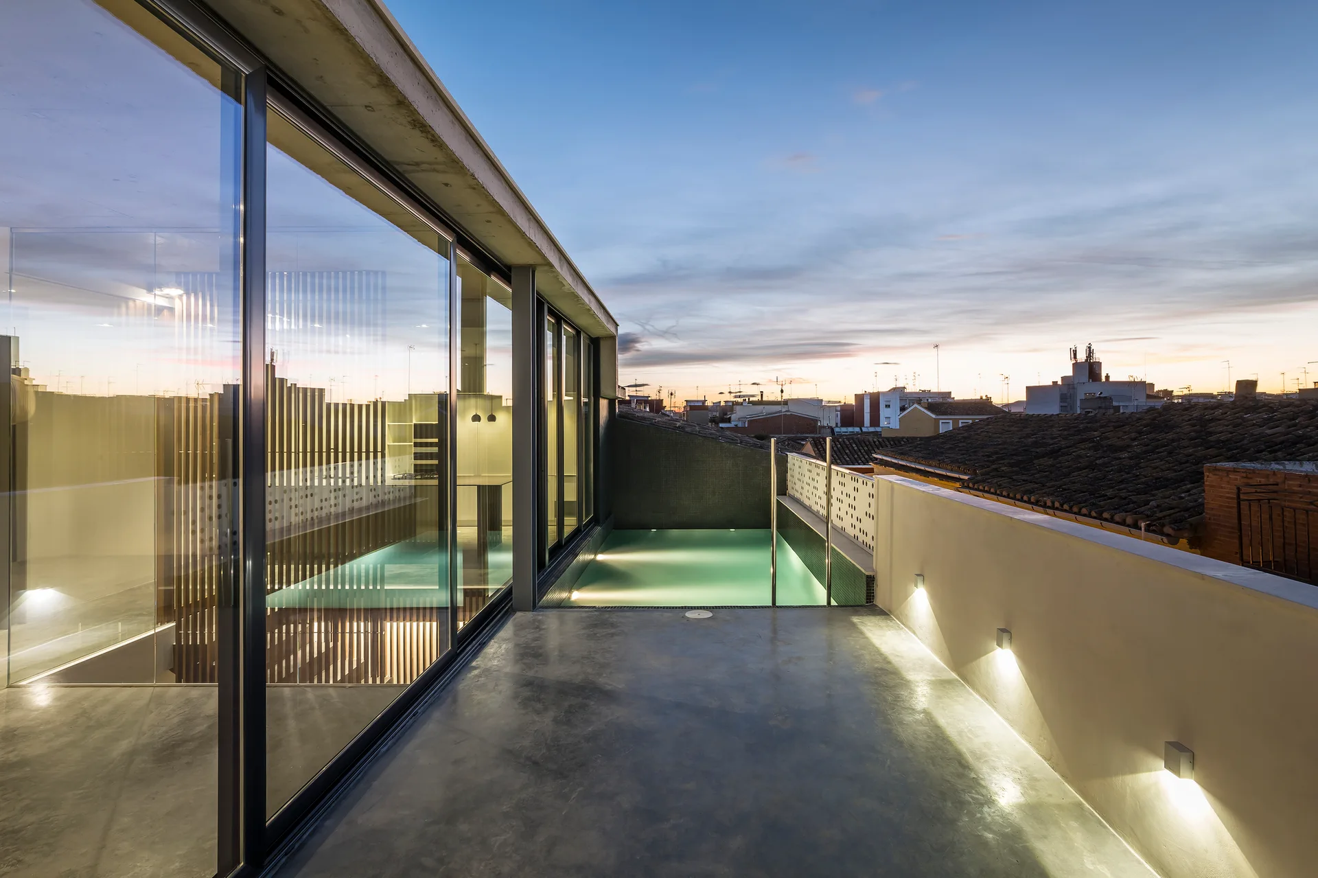 A grey concrete rooftop terrace features a glowing rectangular swimming pool alongside large sliding glass doors and illuminated wall sconces at dusk.
