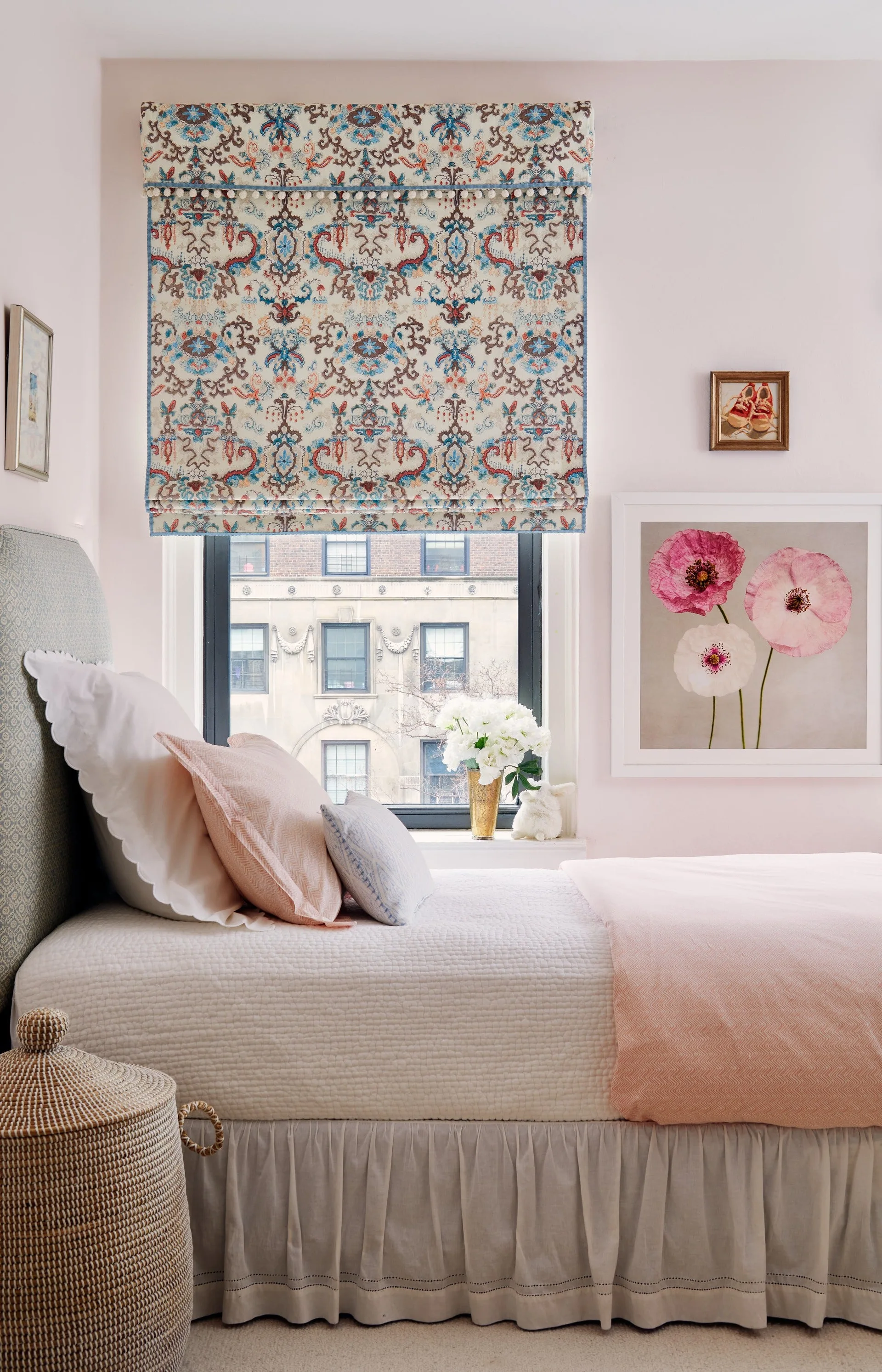 A light pink bedroom features a bed with blush and white pillows, a floral-patterned roman shade over the window, and a framed poppy print on the wall.