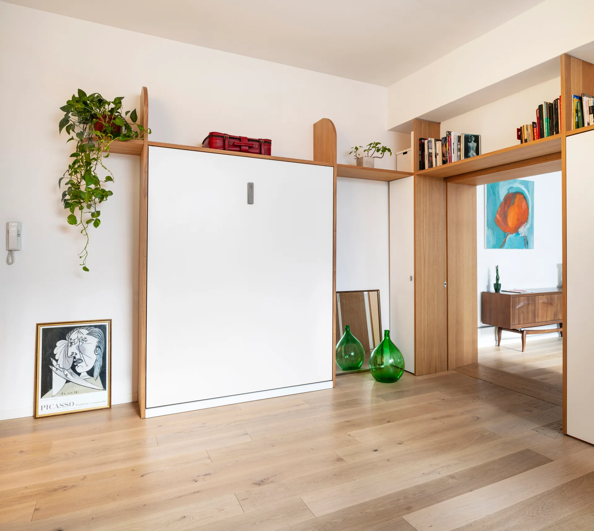 A minimalist room with a white fold-down wall bed built into a light wood frame and archway, featuring green glass bottles on oak floors.