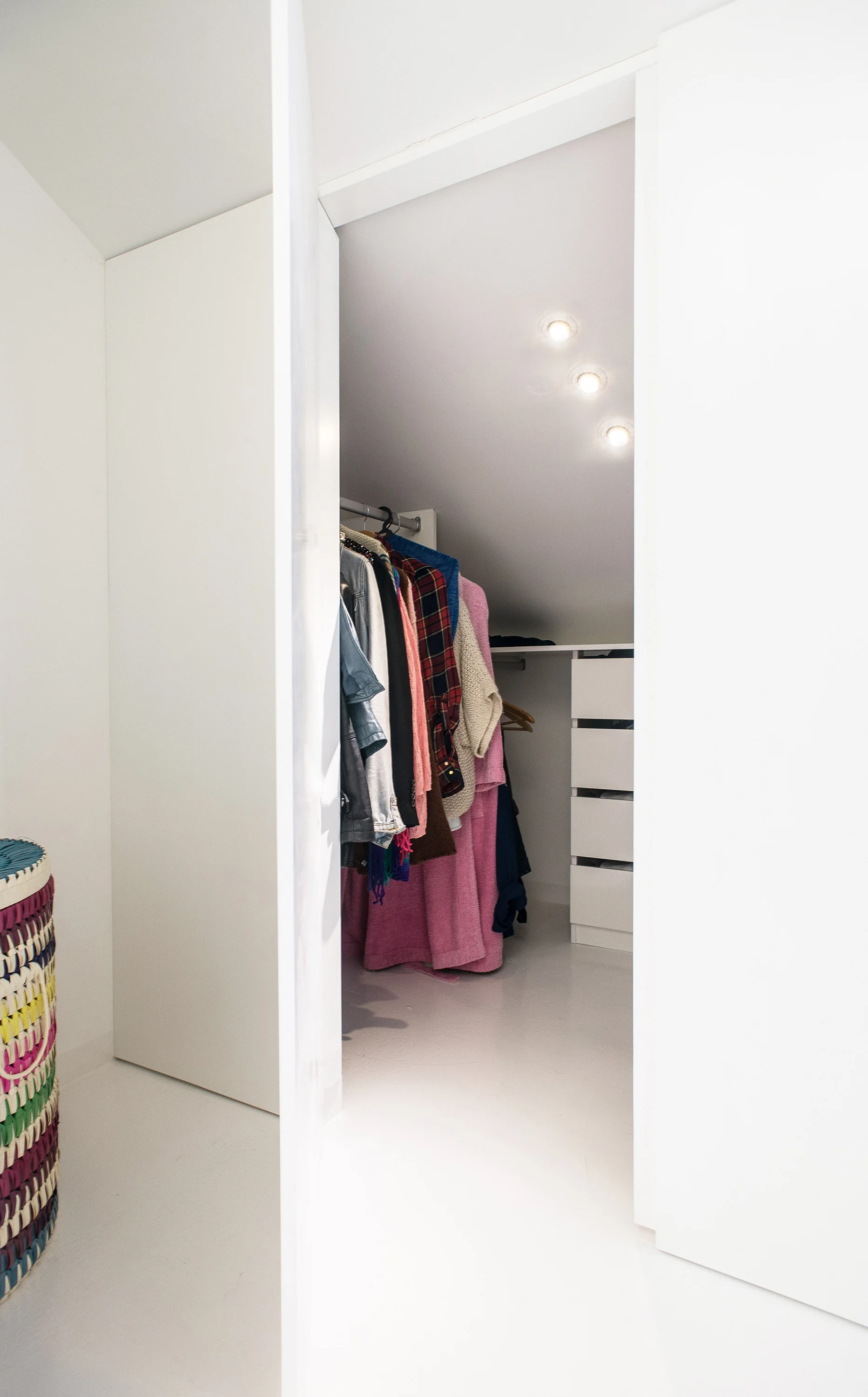 A minimalist white walk-in closet under a sloped ceiling with integrated drawers, hanging clothes on a rod, and recessed ceiling lights.