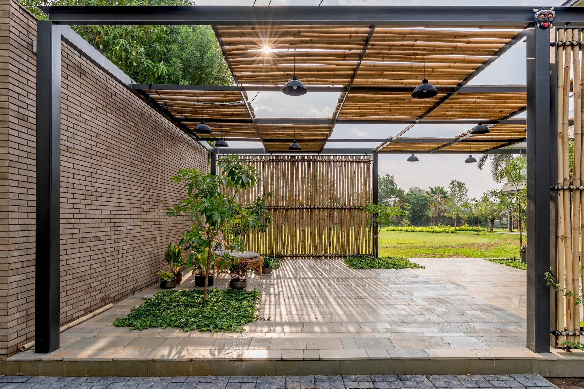 An outdoor patio features a black metal pergola with a bamboo roof, a brown brick wall, stone tile flooring, and integrated green planters.
