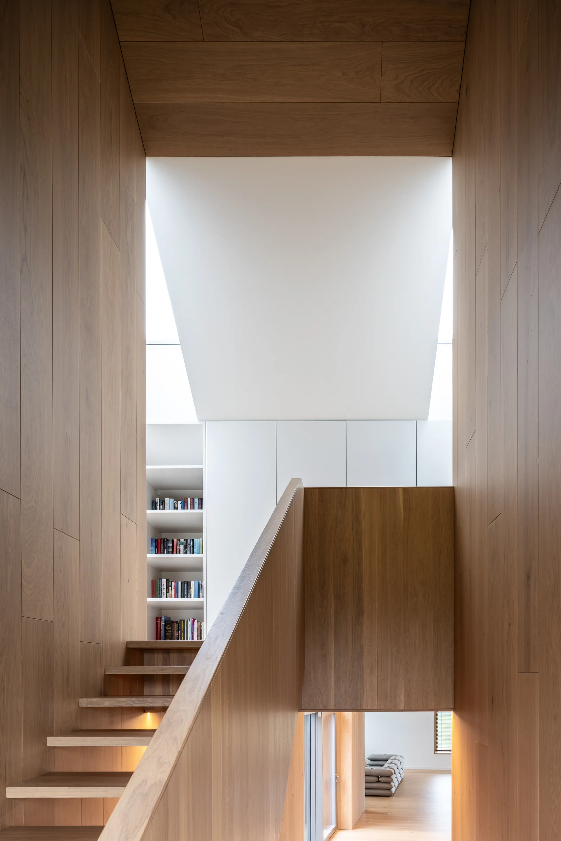 A wooden staircase with under-step lighting features wood-paneled walls and a built-in white bookshelf illuminated by an overhead skylight.