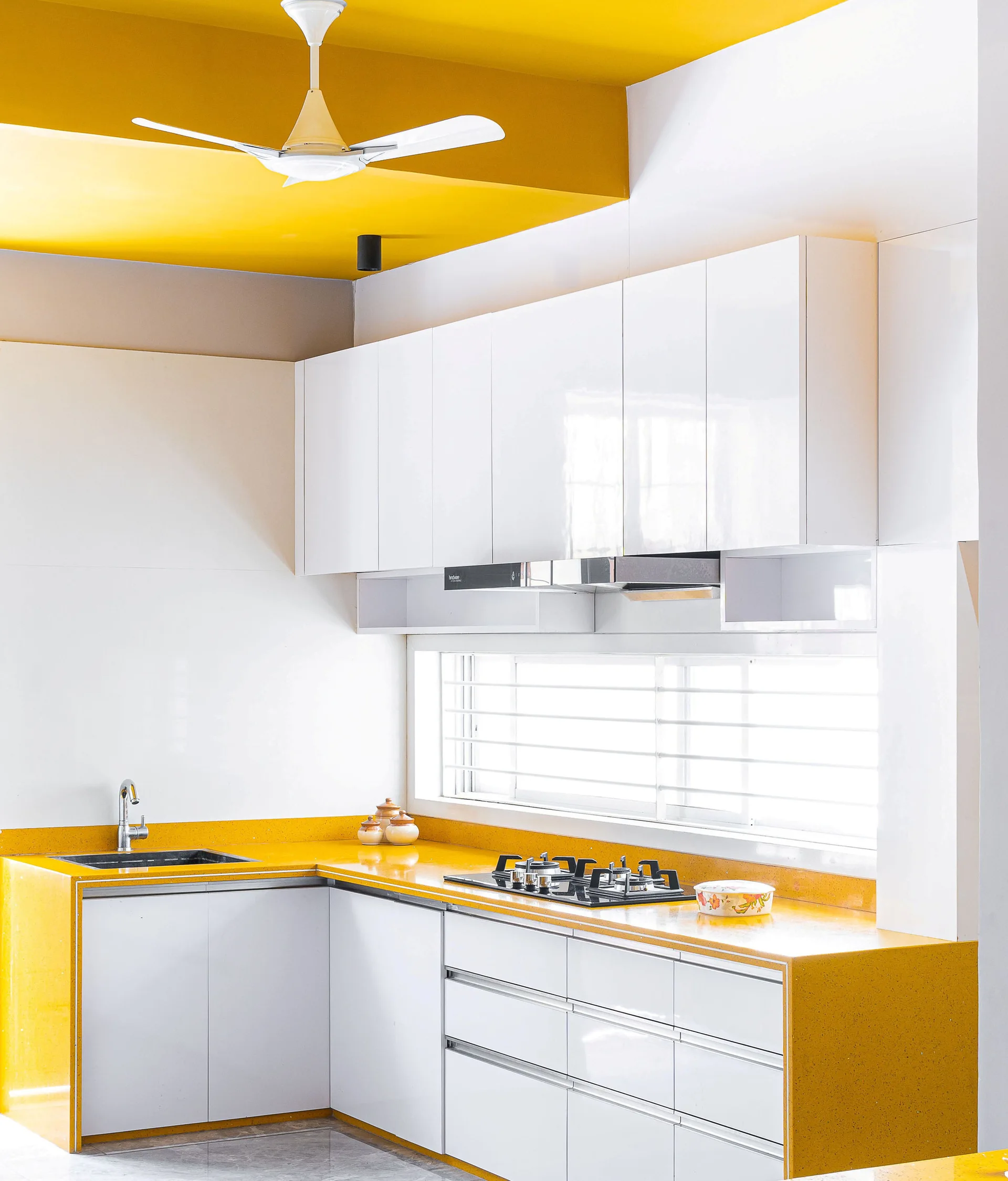 A modern L-shaped kitchen features glossy white cabinets, bold yellow countertops, a built-in cooktop, and a matching yellow ceiling accent.