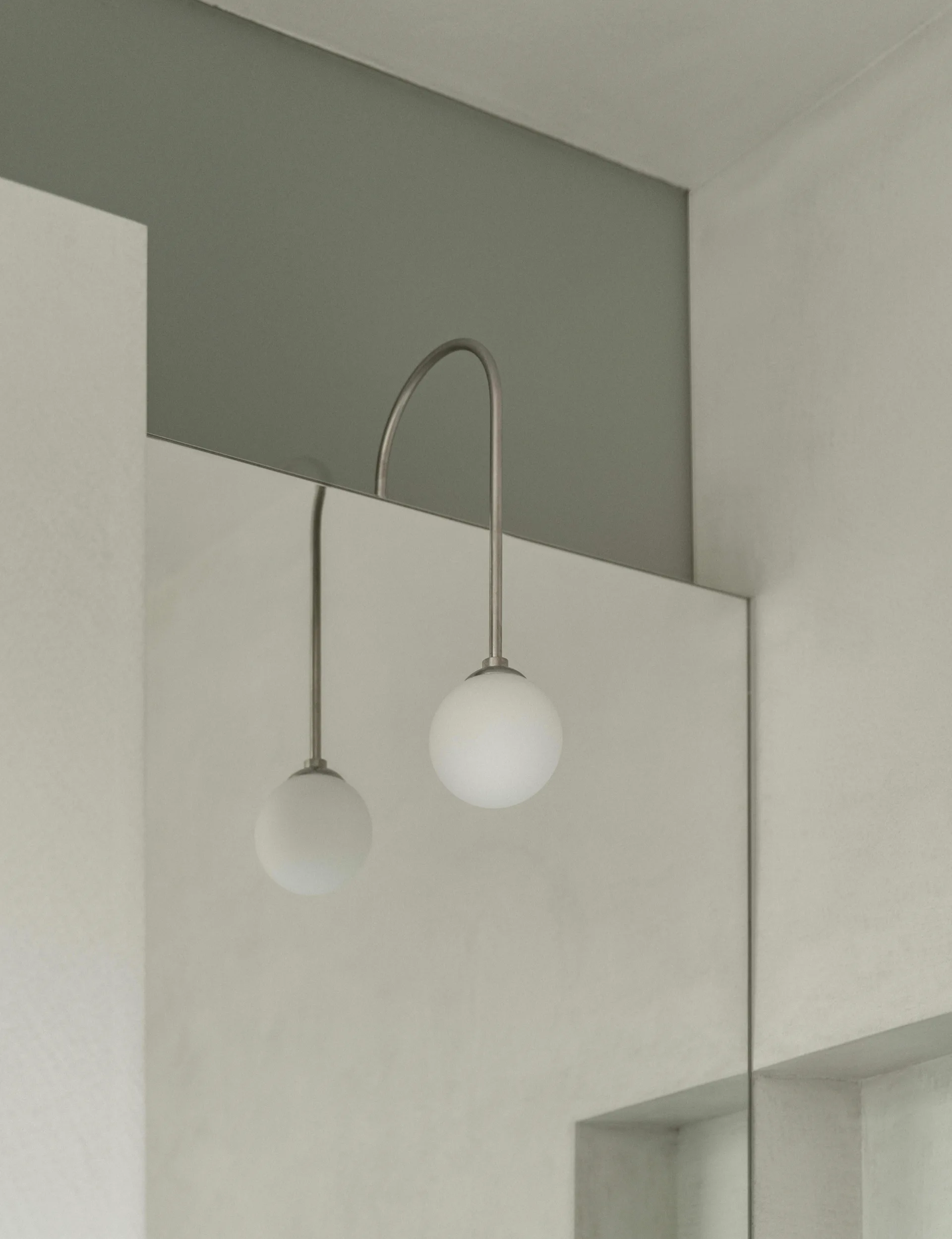 A minimalist wall sconce with a curved metal arm and frosted glass globe hangs over a frameless mirror against a light grey wall.